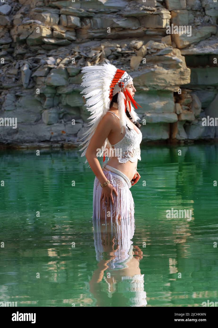 Ein Mädchen wird als Indianerin der Ureinwohner angesehen. Sie ist in Weiß gekleidet und trägt einen gefiederten Kopfschmuck. Man sieht sie in einem Steinbruch-See stehen. Stockfoto