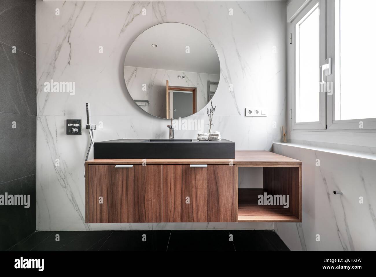 Modernes Design-Badezimmer mit Holzschränken, schwarzem Porzellanwaschbecken, kreisförmigem Spiegelrahmen, Chrommetallarmaturen und -Armaturen sowie schwarzem Marmorboden Stockfoto