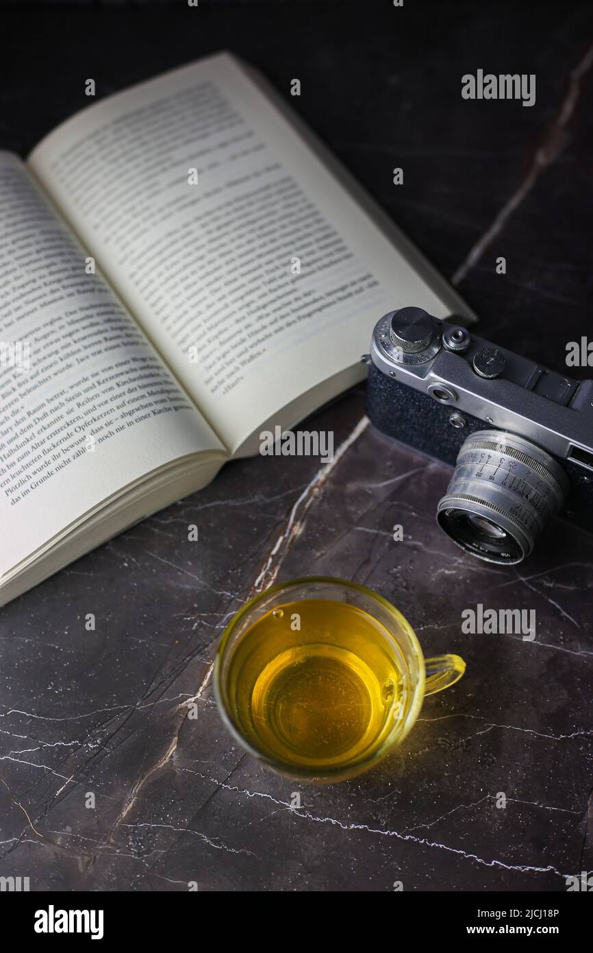 Gebrauter grüner Kräutertee in transparenter Tasse in der Nähe von offenem Buch und Vintage-Kamera auf dunklem Hintergrund Stockfoto Gebrauter grüner Kräutertee in transparenter Tasse in der Nähe von offenem Buch und Vintage-Kamera auf dunklem Hintergrund Stockfoto