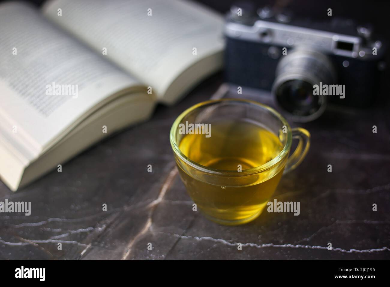 Gebrauter grüner Kräutertee in transparenter Tasse in der Nähe von offenem Buch und Vintage-Kamera auf dunklem Hintergrund Stockfoto Gebrauter grüner Kräutertee in transparenter Tasse in der Nähe von offenem Buch und Vintage-Kamera auf dunklem Hintergrund Stockfoto