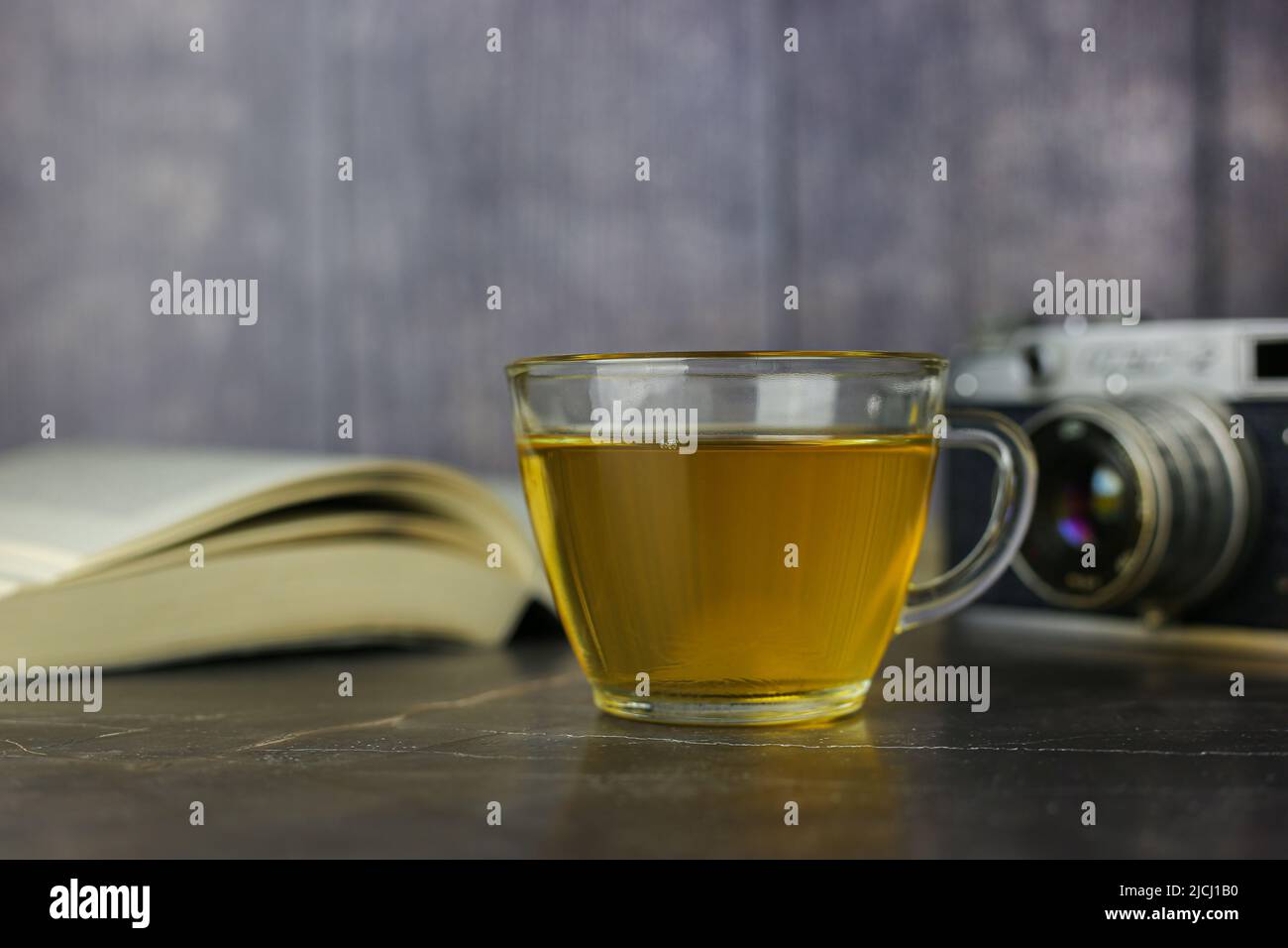 Gebrauter grüner Kräutertee in transparenter Tasse in der Nähe von offenem Buch und Vintage-Kamera auf dunklem Hintergrund Stockfoto Gebrauter grüner Kräutertee in transparenter Tasse in der Nähe von offenem Buch und Vintage-Kamera auf dunklem Hintergrund Stockfoto