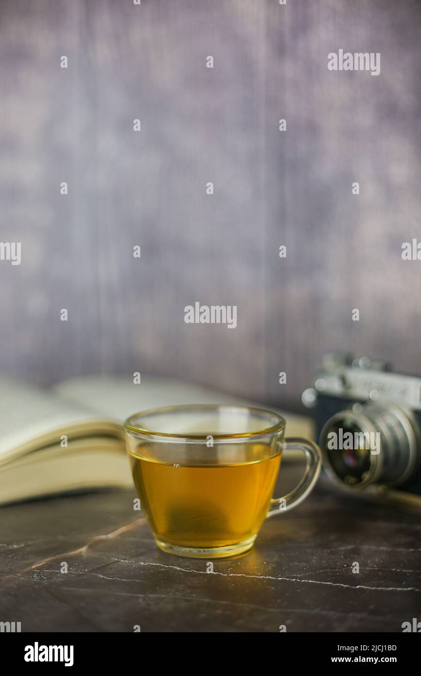 Gebrauter grüner Kräutertee in transparenter Tasse in der Nähe von offenem Buch und Vintage-Kamera auf dunklem Hintergrund Stockfoto Gebrauter grüner Kräutertee in transparenter Tasse in der Nähe von offenem Buch und Vintage-Kamera auf dunklem Hintergrund Stockfoto