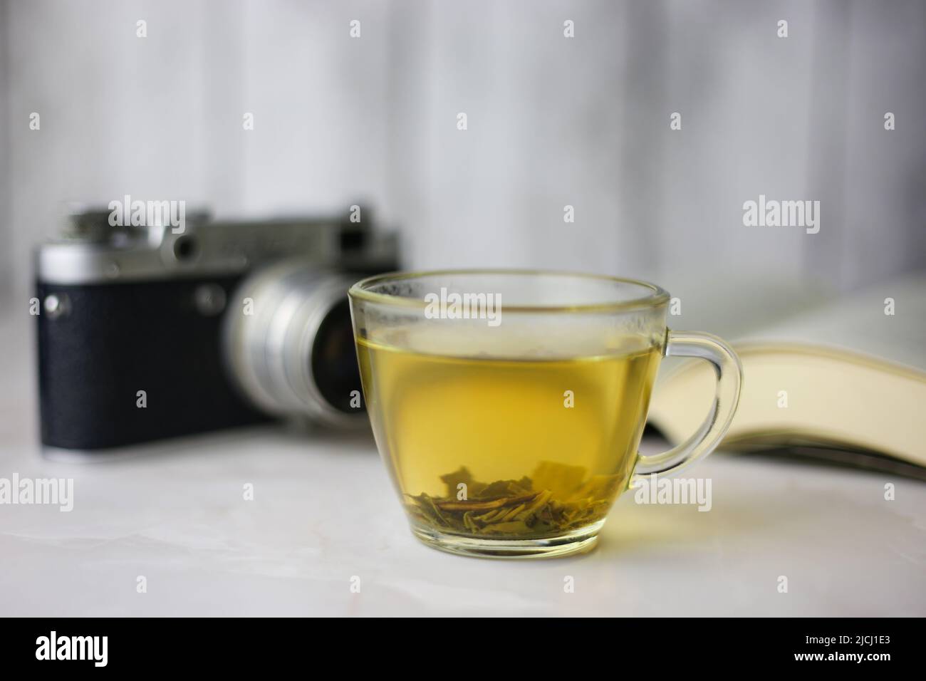 Gebrauter grüner Kräutertee in einer transparenten Tasse neben einem offenen Buch und einer Vintage-Kamera auf hellem Hintergrund Stockfoto Gebrauter grüner Kräutertee in einer transparenten Tasse neben einem offenen Buch und einer Vintage-Kamera auf hellem Hintergrund Stockfoto