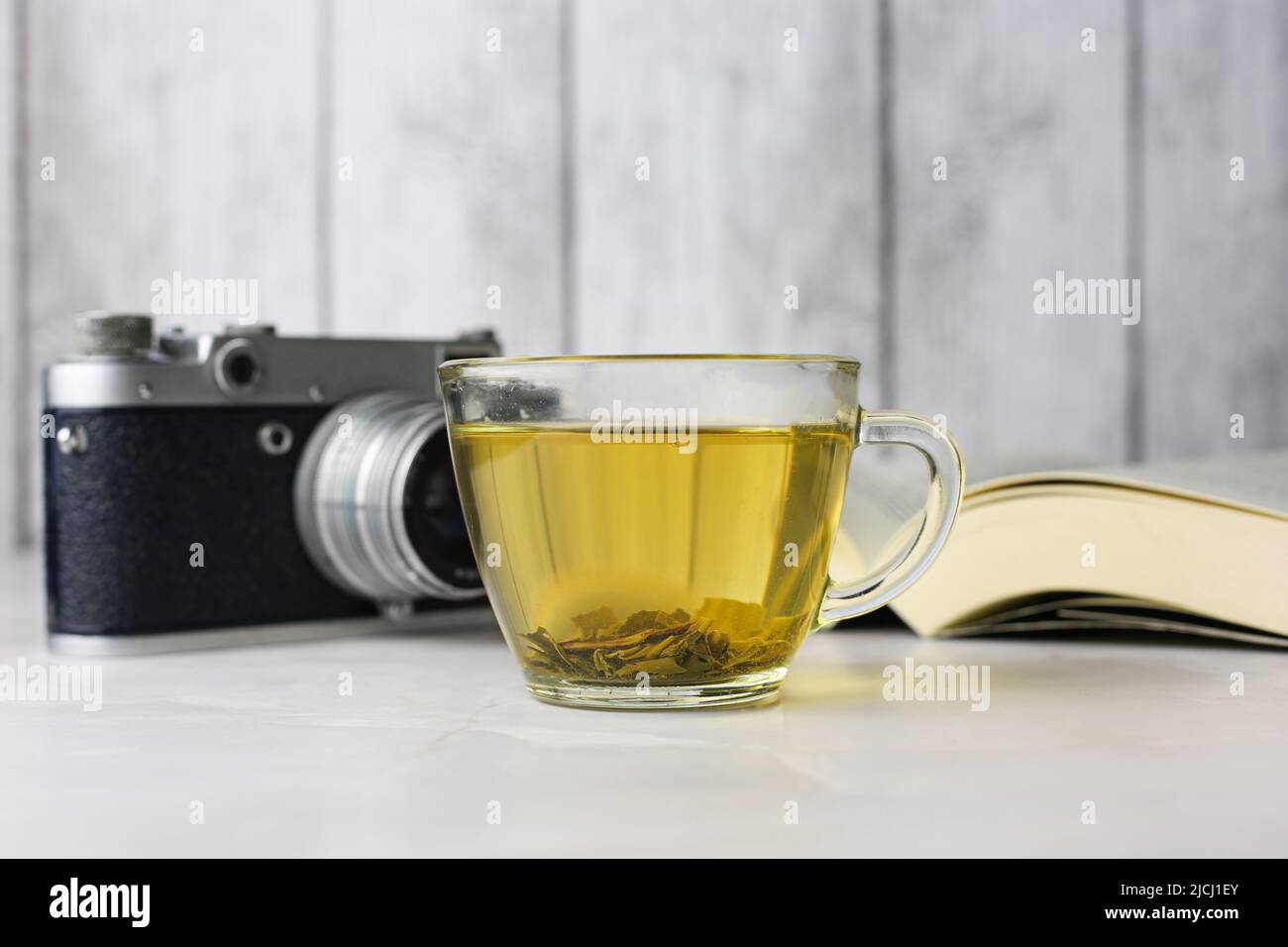 Gebrauter grüner Kräutertee in einer transparenten Tasse neben einem offenen Buch und einer Vintage-Kamera auf hellem Hintergrund Stockfoto Gebrauter grüner Kräutertee in einer transparenten Tasse neben einem offenen Buch und einer Vintage-Kamera auf hellem Hintergrund Stockfoto