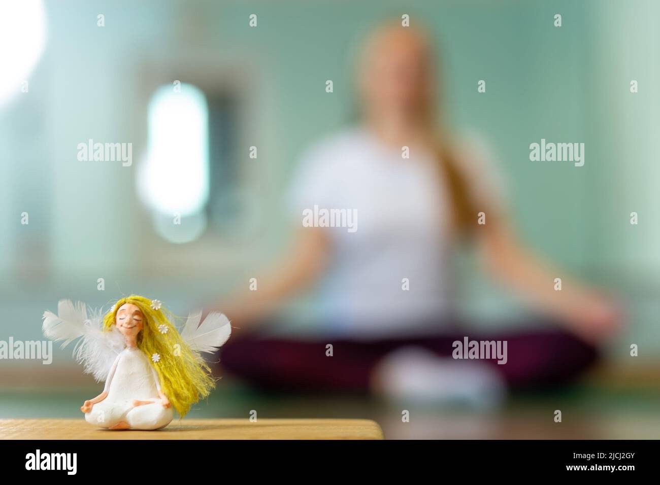 Eine Puppe in Form eines Engels auf unscharfem Hintergrund mit gesunder Frau, die drinnen Yoga praktiziert. Die Yoga-Anfängerin ist in Lotushaltung. Die Position r Stockfoto
