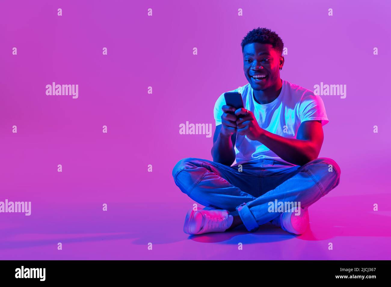 Afroamerikanischer Typ sitzt mit gekreuzten Beinen mit dem Smartphone, surft in sozialen Medien, chattet mit einem Freund im Neonlicht Stockfoto