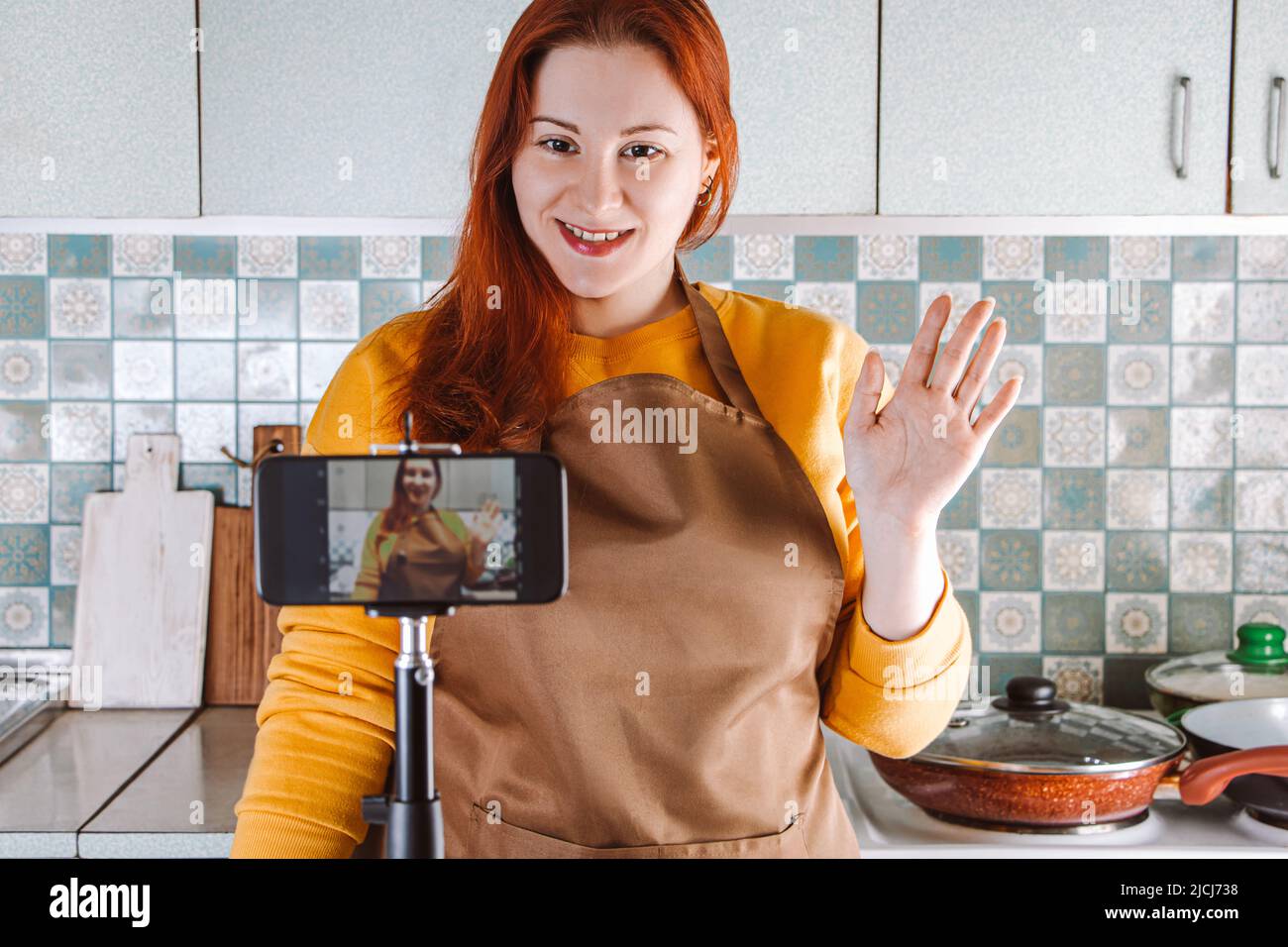 Moderne Frau Blogger ist Live-Streaming und Aufnahme Video Rezept für Lebensmittel-Blog. Junge rothaarige Frau in gelber Schürze Kochen Lebensmittel online in Home KI Stockfoto