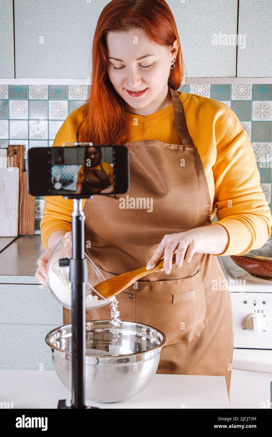Moderne Frau Blogger ist Live-Streaming und Aufnahme Video Rezept für Lebensmittel-Blog. Junge rothaarige Frau in gelber Schürze Kochen Lebensmittel online in Home KI Stockfoto