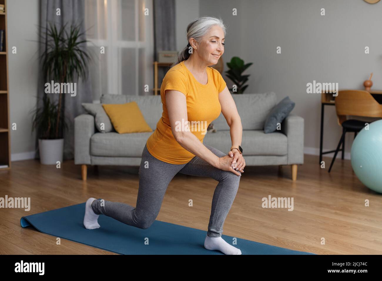 Positive sportliche Senior-Frau, die sich zu Hause fit hält, Ausfallschritte macht, im Wohnzimmer auf einer Yogamatte trainiert und lächelt Stockfoto