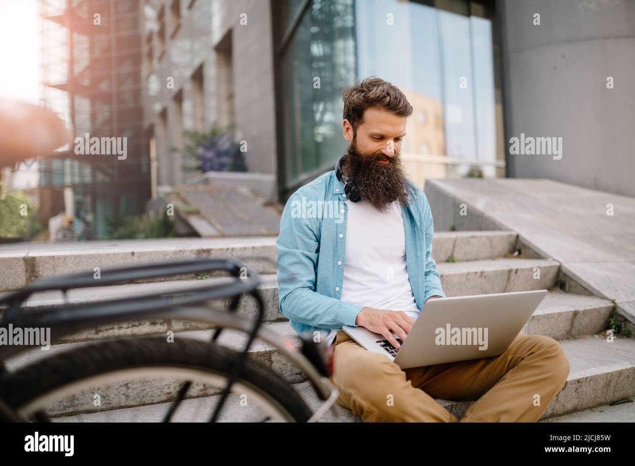 Bartmann Unternehmer sitzt draußen, arbeitet an seinem Laptop. Er ist entspannt, lächelt und genießt seine Zeit draußen. Stockfoto