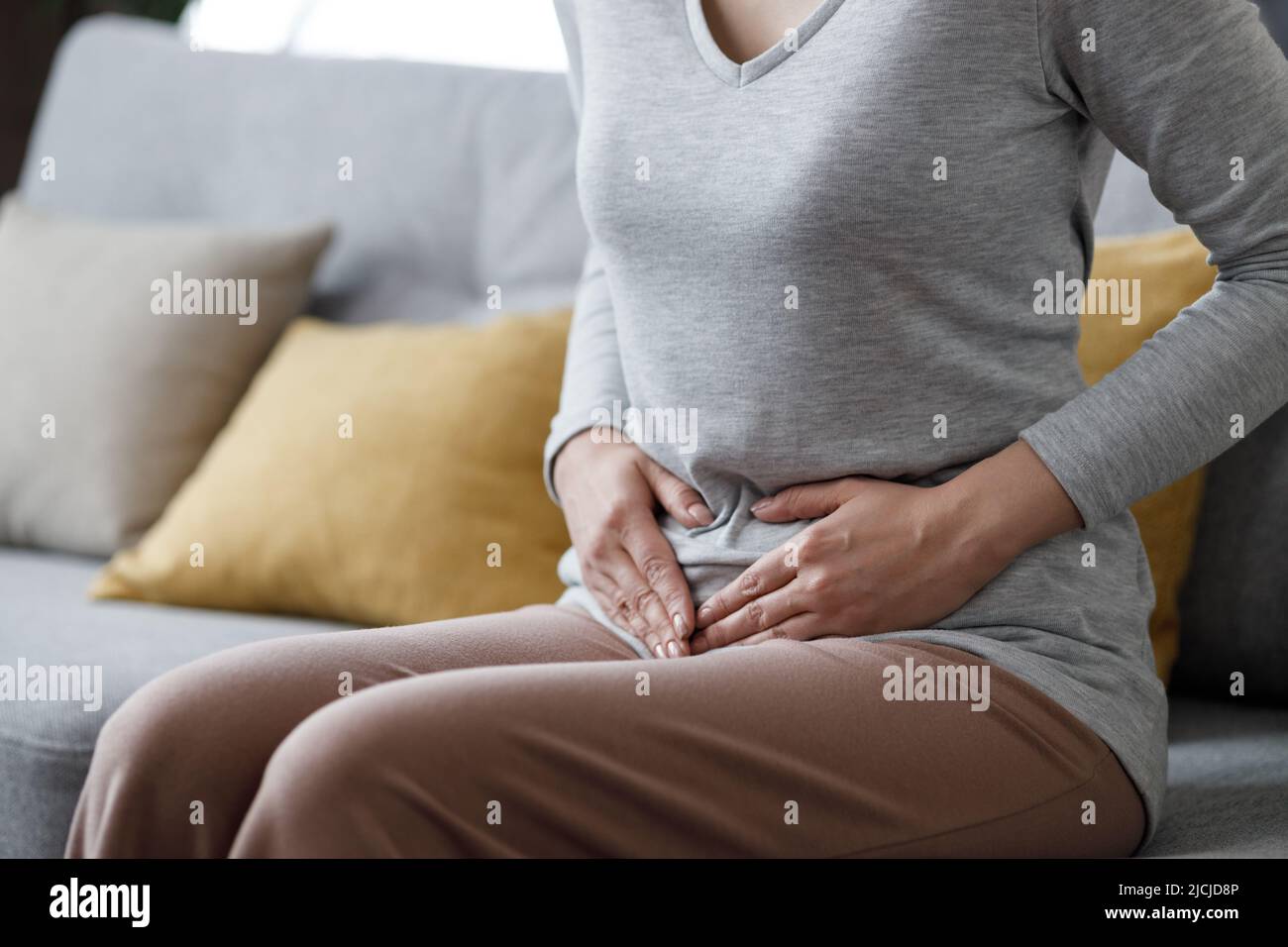 Kranke junge Frau, die an Menstruationsschmerzen leidet. Frau mit den Händen, die den Bauch quetschen, mit schmerzhaften Bauchschmerzen oder Periodenkrämpfen, die auf dem Sofa sitzen Stockfoto