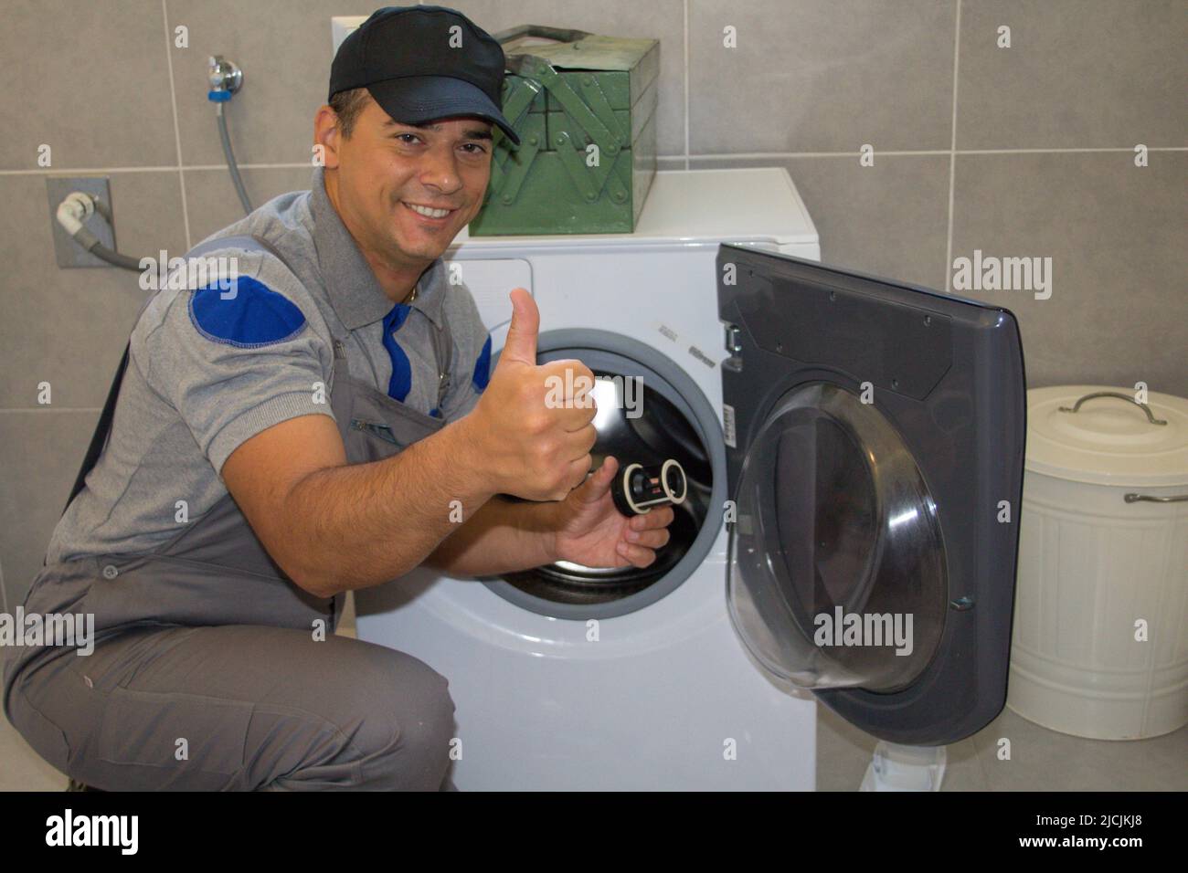 Foto eines lächelnden Klempners mit Daumen nach oben, als er eine defekte Waschmaschine repariert. Stockfoto