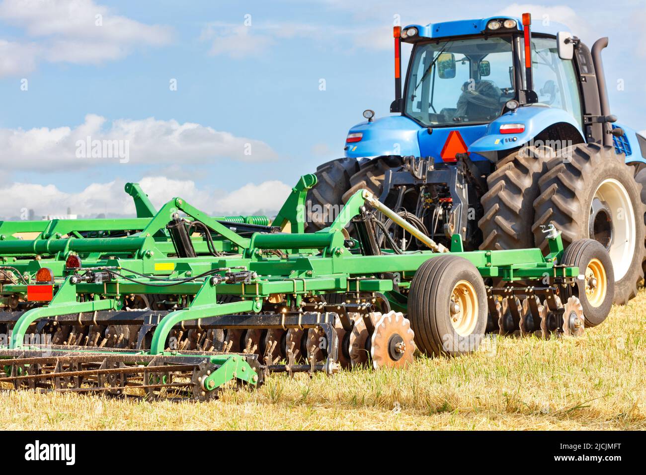 Eine Scheibenegge klammert sich an einen landwirtschaftlichen Traktor auf Rädern, um Land auf den Feldern vor einem blauen Himmel mit weißen Wolken zu kultivieren. Speicherplatz kopieren. Stockfoto