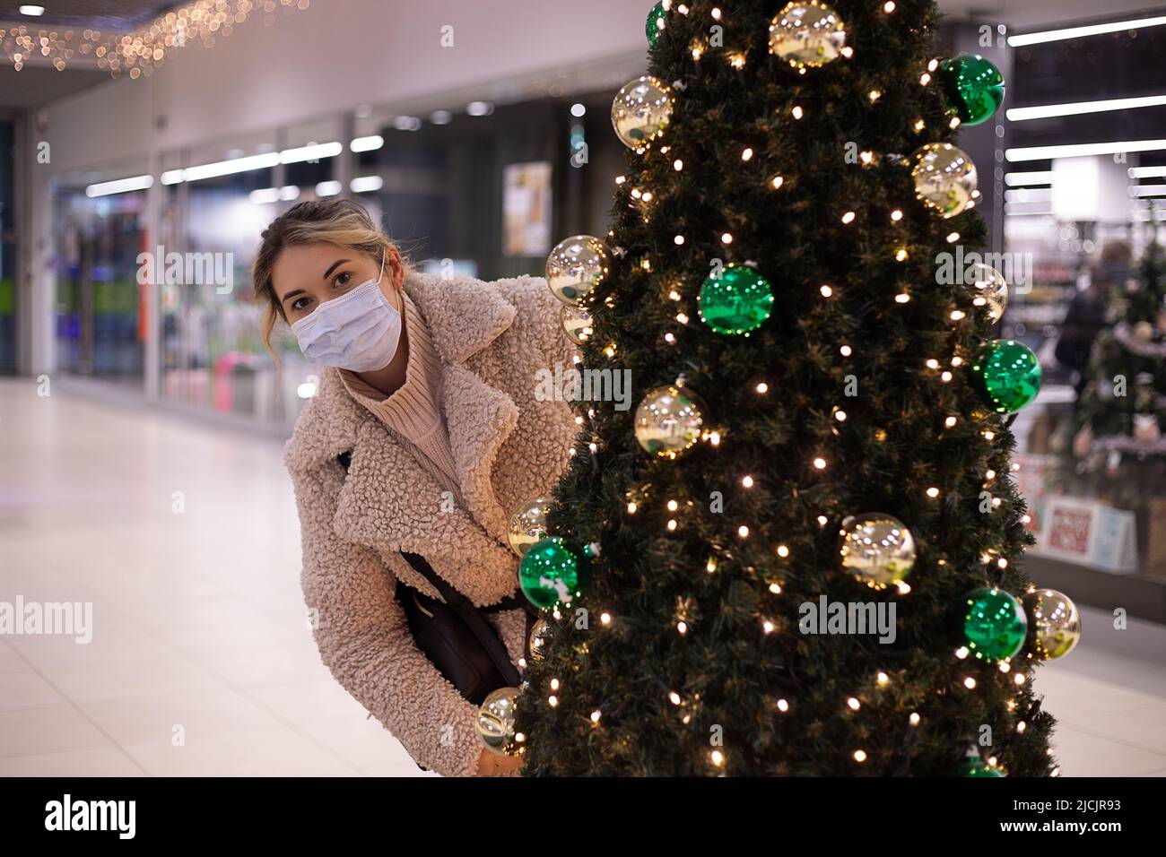 Nette Frau mit Spaß Blick aus hinter geschmückten Weihnachtsbaum im Einkaufszentrum. Frau in medizinischer Maske. Einkaufszentrum zu Weihnachten. Neuer Normalwert Stockfoto