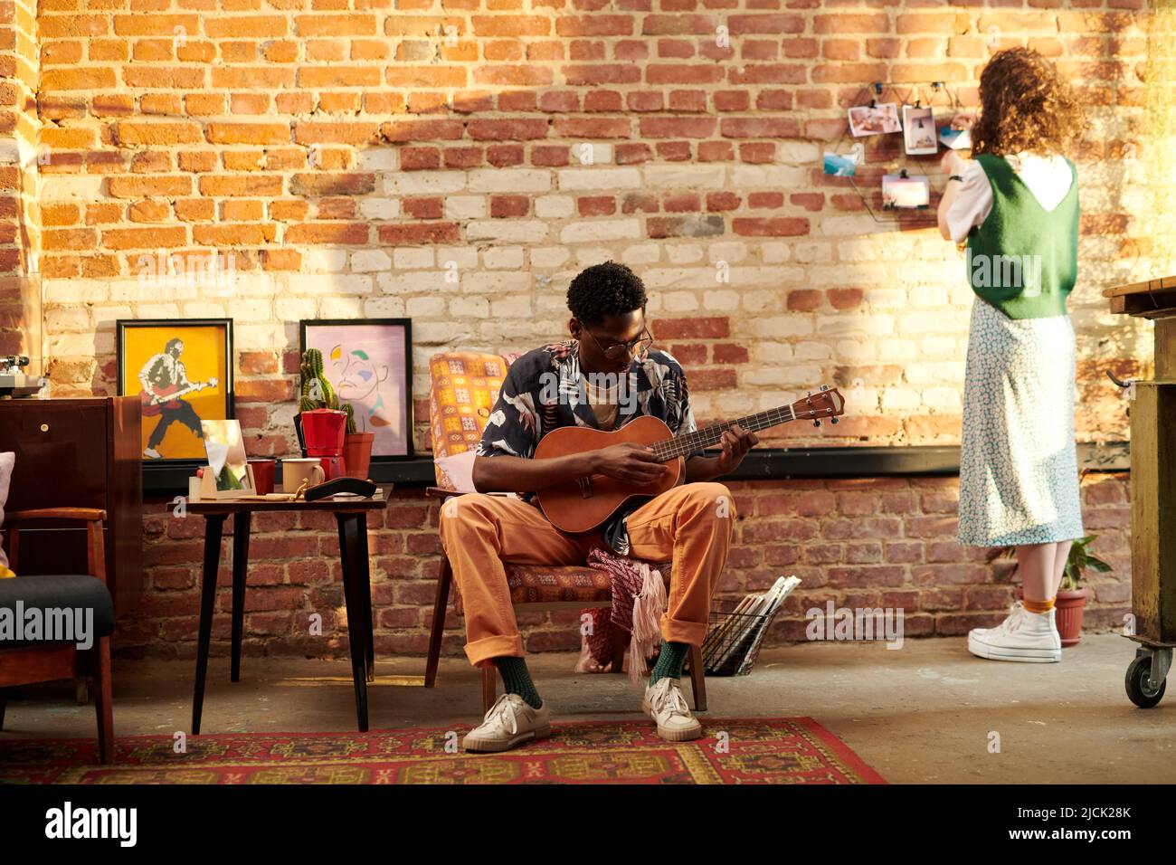 Junger Mann, der im Sessel sitzt und akustische Gitarre spielt, während seine Freundin vor einer Backsteinmauer steht und Fotos anschaut Stockfoto