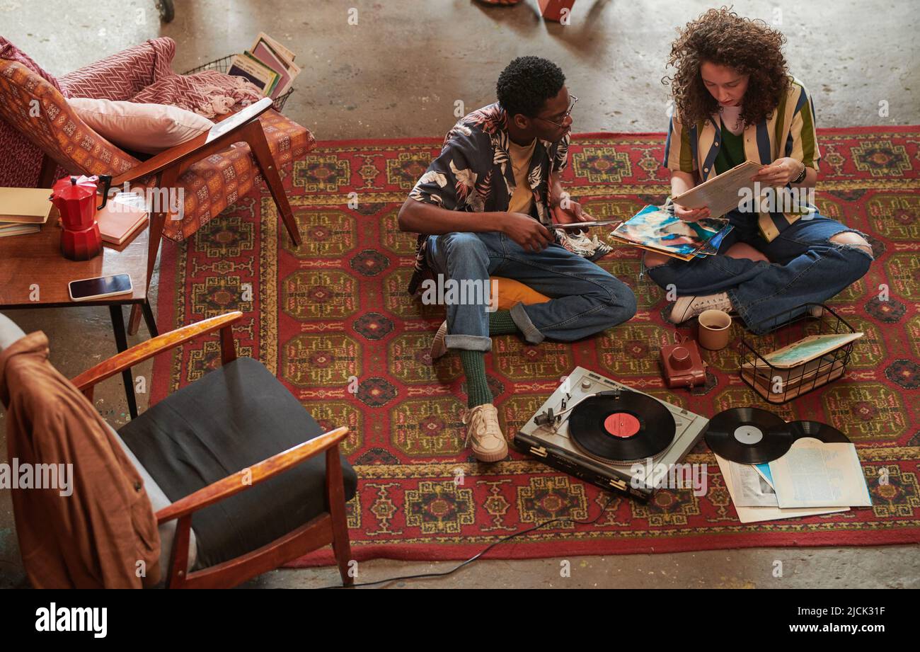 Eine junge Frau zeigt ihrem afroamerikanischen Freund ihre Lieblingsvynil-Scheibe in stilvoller Casualwear, die neben ihr auf dem roten Teppich sitzt Stockfoto