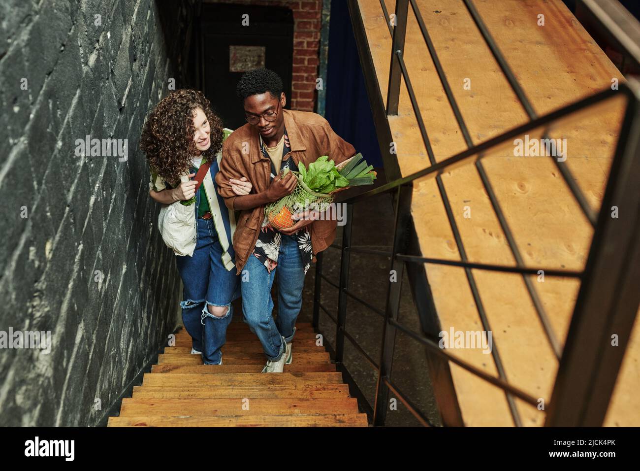 Junge interracial Paar in Casualwear bewegen sich nach oben auf Holztreppe in Loft-Wohnung, während sie aus dem Supermarkt zurück Stockfoto
