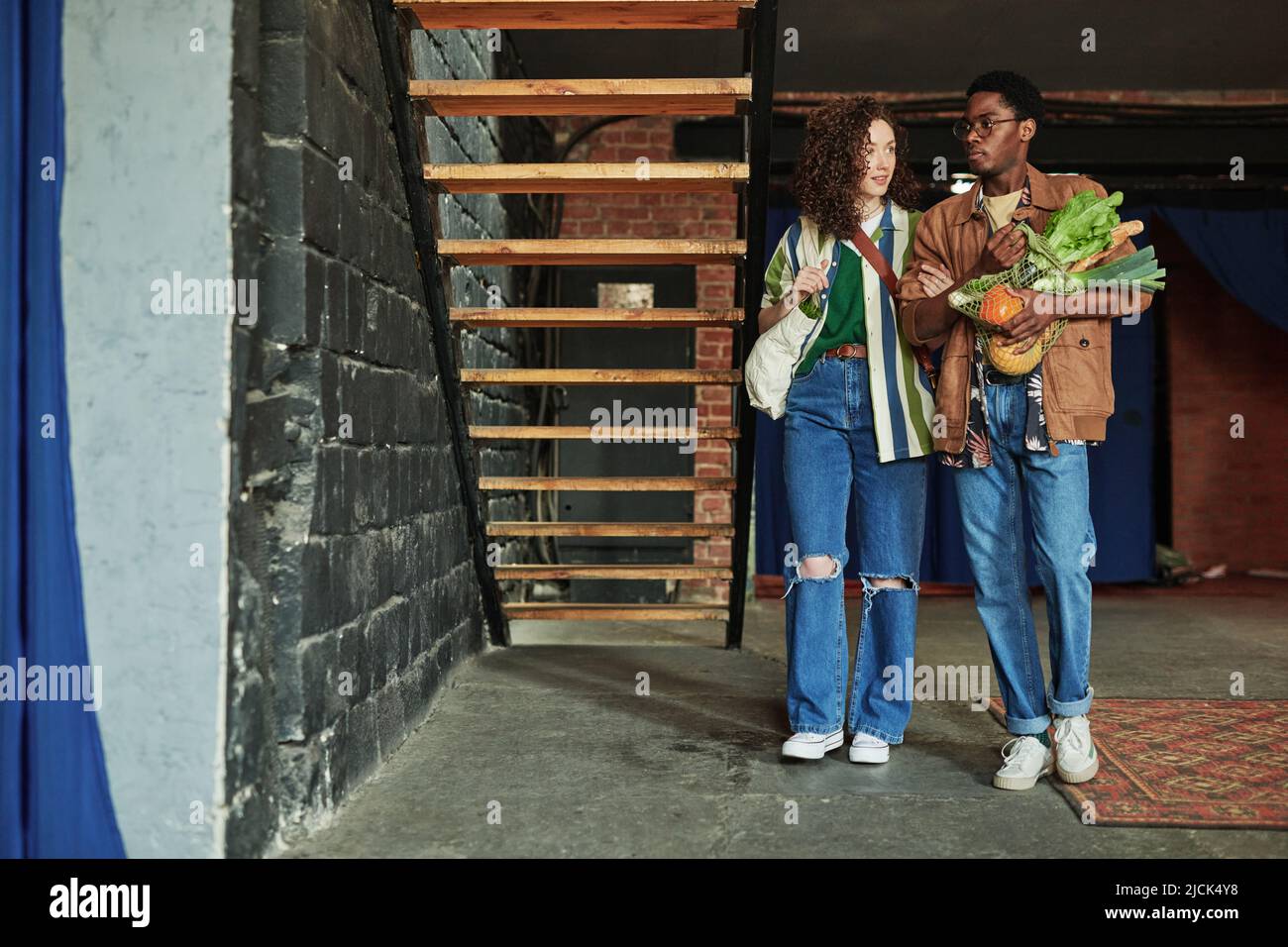 Junge Frau hält ihren Freund mit frischem Gemüse in der Einkaufstasche am Arm, während beide sich in ihrer Loft-Wohnung bewegen Stockfoto