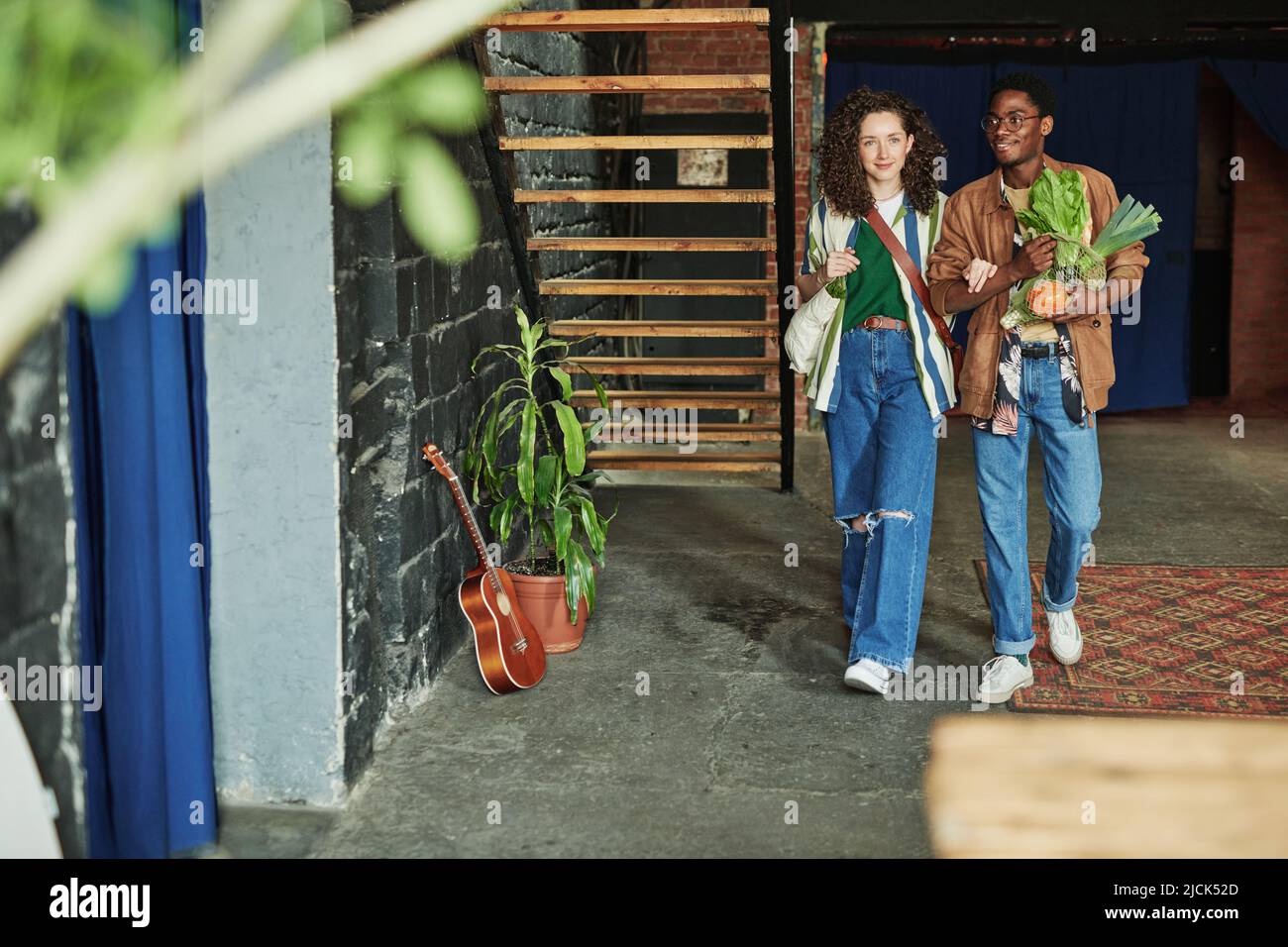 Glückliches junges interkulturelles Paar in stilvoller Casualwear, das sich im Wohnzimmer der Loft-Wohnung bewegt, während es vom Supermarkt zurückkommt Stockfoto