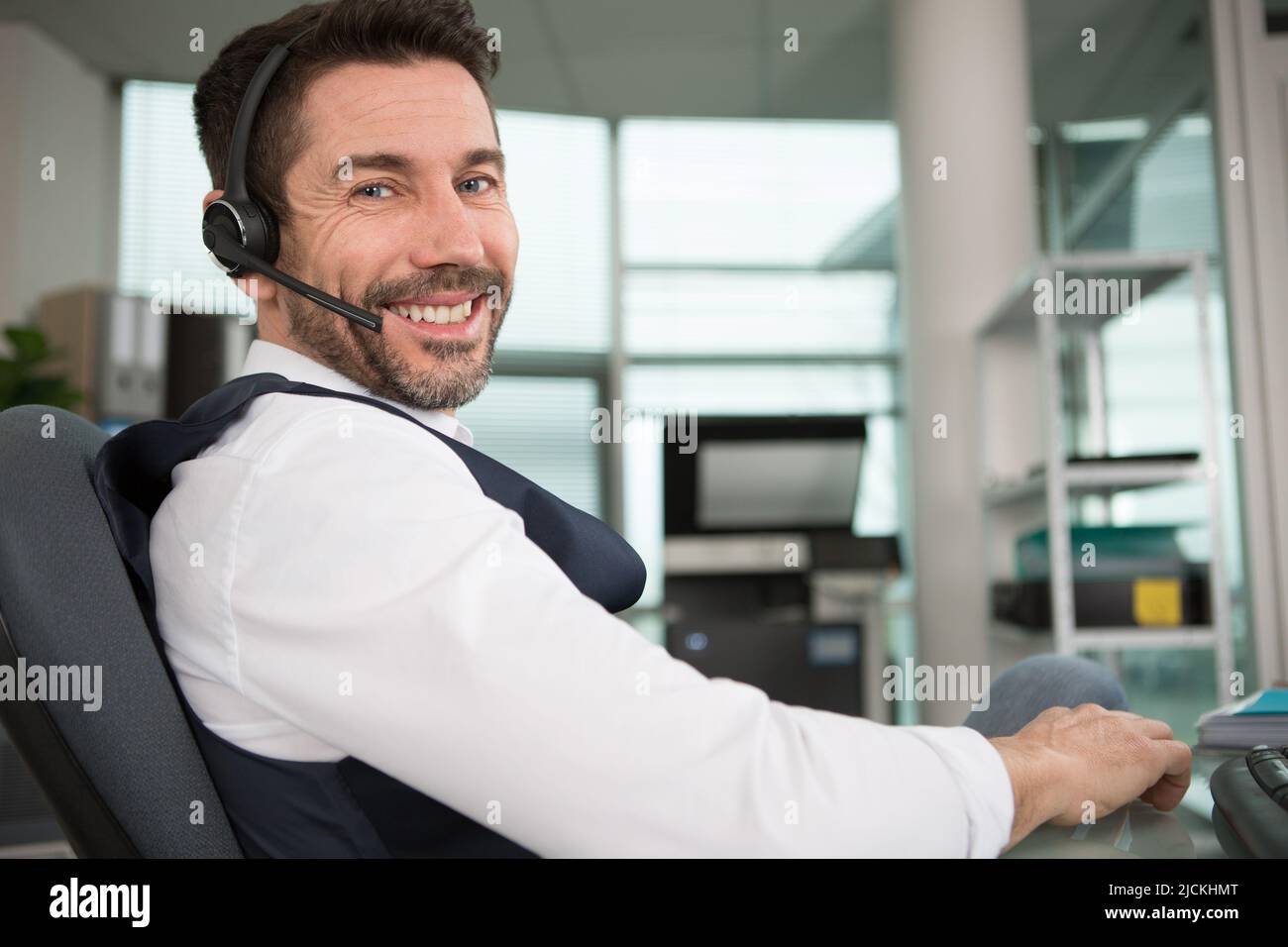 Ein Geschäftsmann, der in einem vielbeschäftigten Telefonbüro ein Headset trägt, lächelt der Kamera zu Stockfoto
