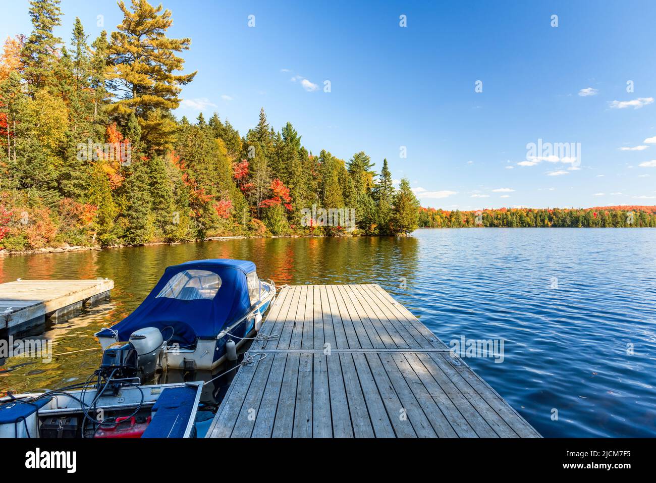 Motorboote wurden an einem hölzernen Steg an einem See mit bewaldeten Ufern an einem sonnigen Herbsttag auf der Spitze des Herbstlaubes befestigt Stockfoto