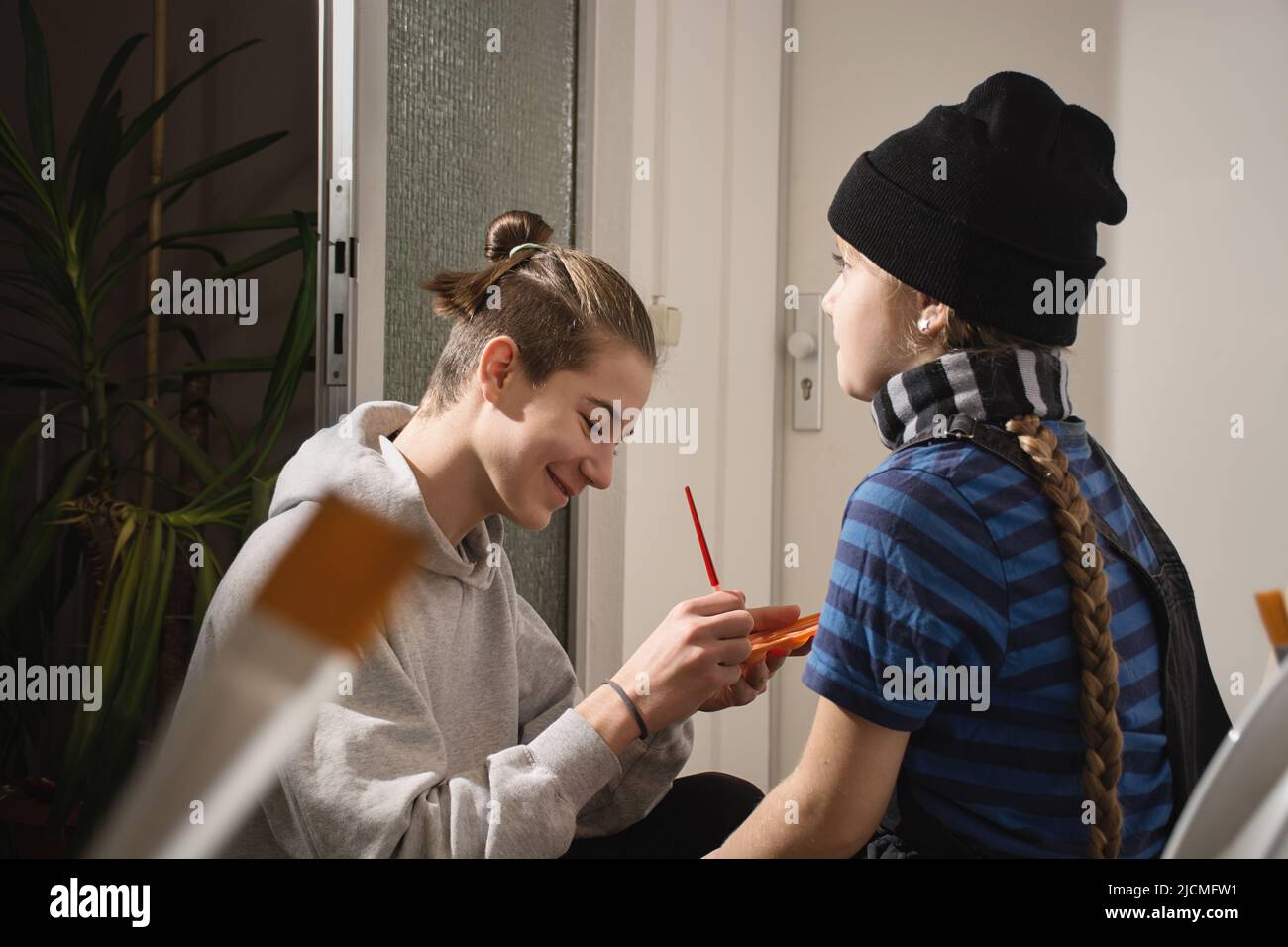 Der Teenager zeichnet zu Hause einen Pinsel für Gesichtszeichnungen auf das Gesicht seiner jüngeren Schwester. Hut und Schal auf Mädchen. Alternatives Bild für Halloween, Maskeraden, Geburtstag. Echte Menschen. Defokussieren Sie den Vordergrund. Stockfoto