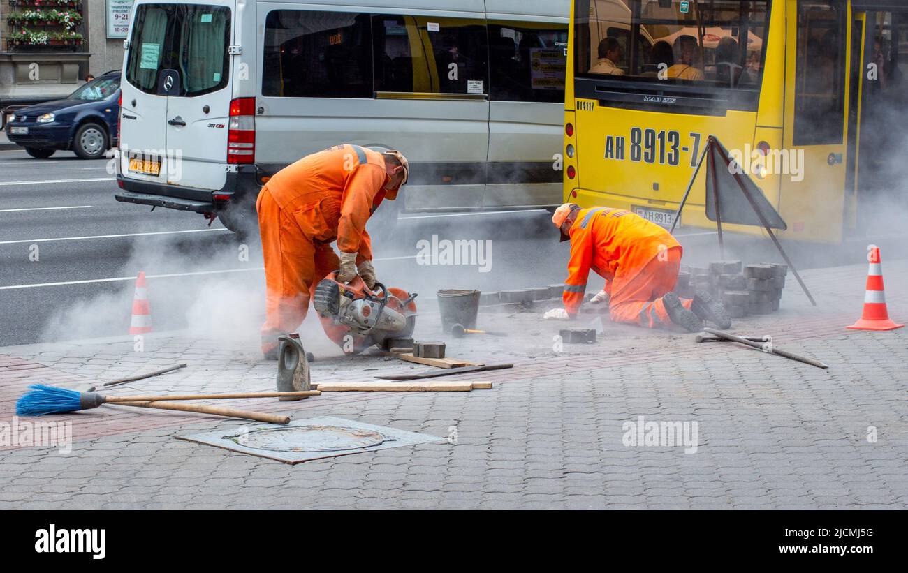 Straßenarbeiter legen Pflasterplatten. Eine mit einer Kreissäge schneidet Betonblöcke. Stockfoto