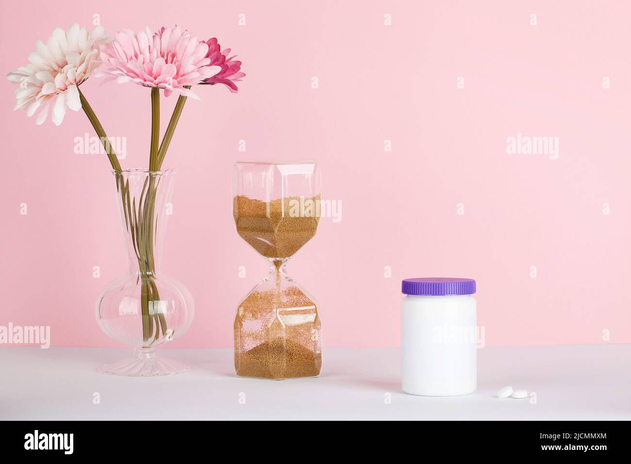 Frauen Gesundheit, mittleres Alter und Wechseljahre Konzept. Gesundheitsstil. Copy space. Weiße Plastikflasche mit Pillen, Blumen, Sanduhr auf rosa Hintergrund Stockfoto