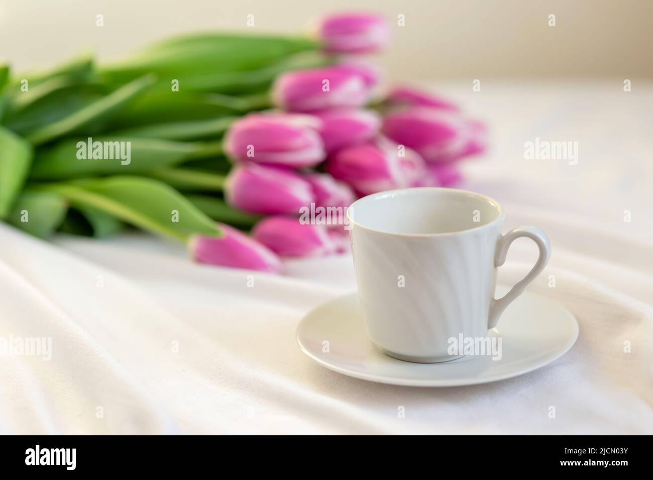Auf einer weißen Tischdecke liegt eine weiße Tasse mit einer Untertasse und einem Bouquet aus schönen rosa Tulpen. Stockfoto