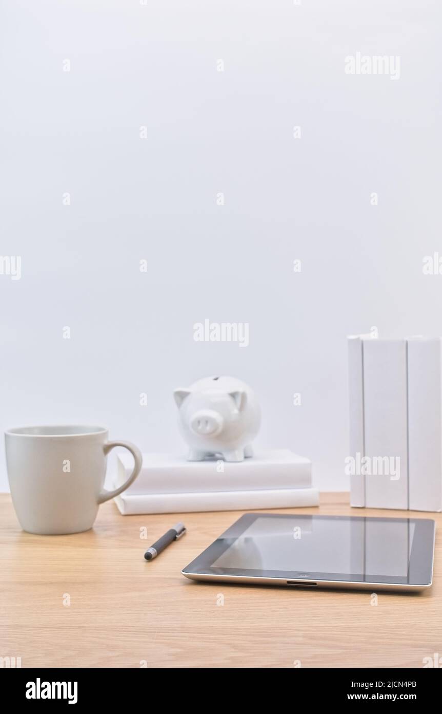 Apple ipad Tablet mit Büchern Stift Becher und Sparschwein auf einem hellen Holzschreibtisch gegen weiße Wand Stockfoto