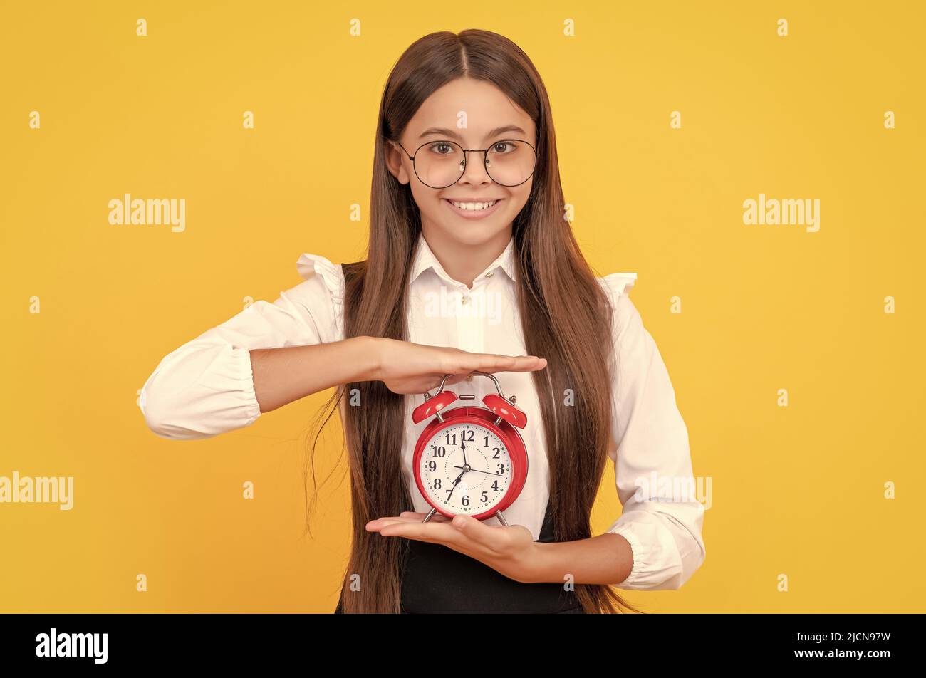 Fröhliches Kind in Schuluniform und Brille mit Wecker, der die Zeit und die Schulzeit anzeigt. Stockfoto