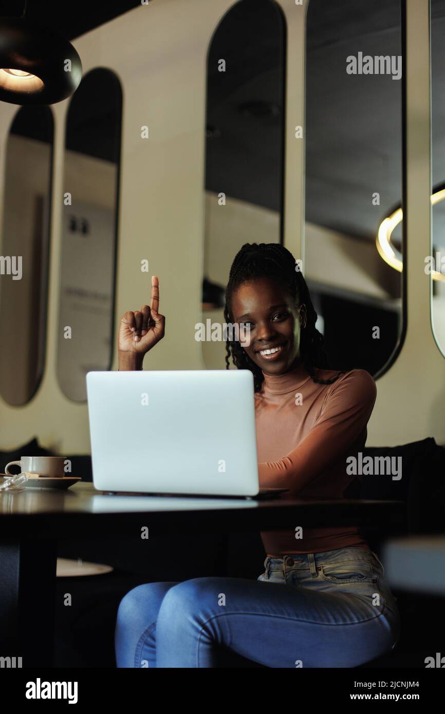 Vertikale multikulturelle dunkle Haut Frau trinken Kaffee und arbeiten mit Laptop, um Daten im Café zu analysieren. Zeigefinger nach oben zeigen Stockfoto