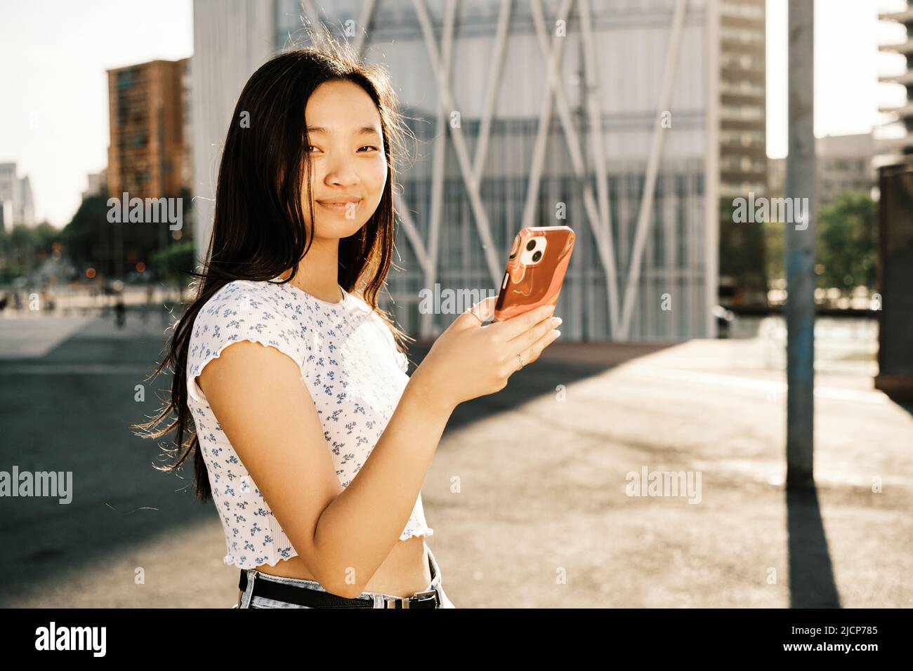 Junge asiatische Frau, die auf die Kamera schaut, während sie ihr Mobiltelefon auf der Straße im Freien benutzt. Stockfoto