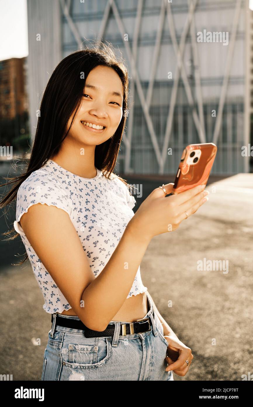 Junge asiatische Frau schaut auf die Kamera und lächelt, während sie ein Mobiltelefon im Freien auf der Straße hält. Stockfoto