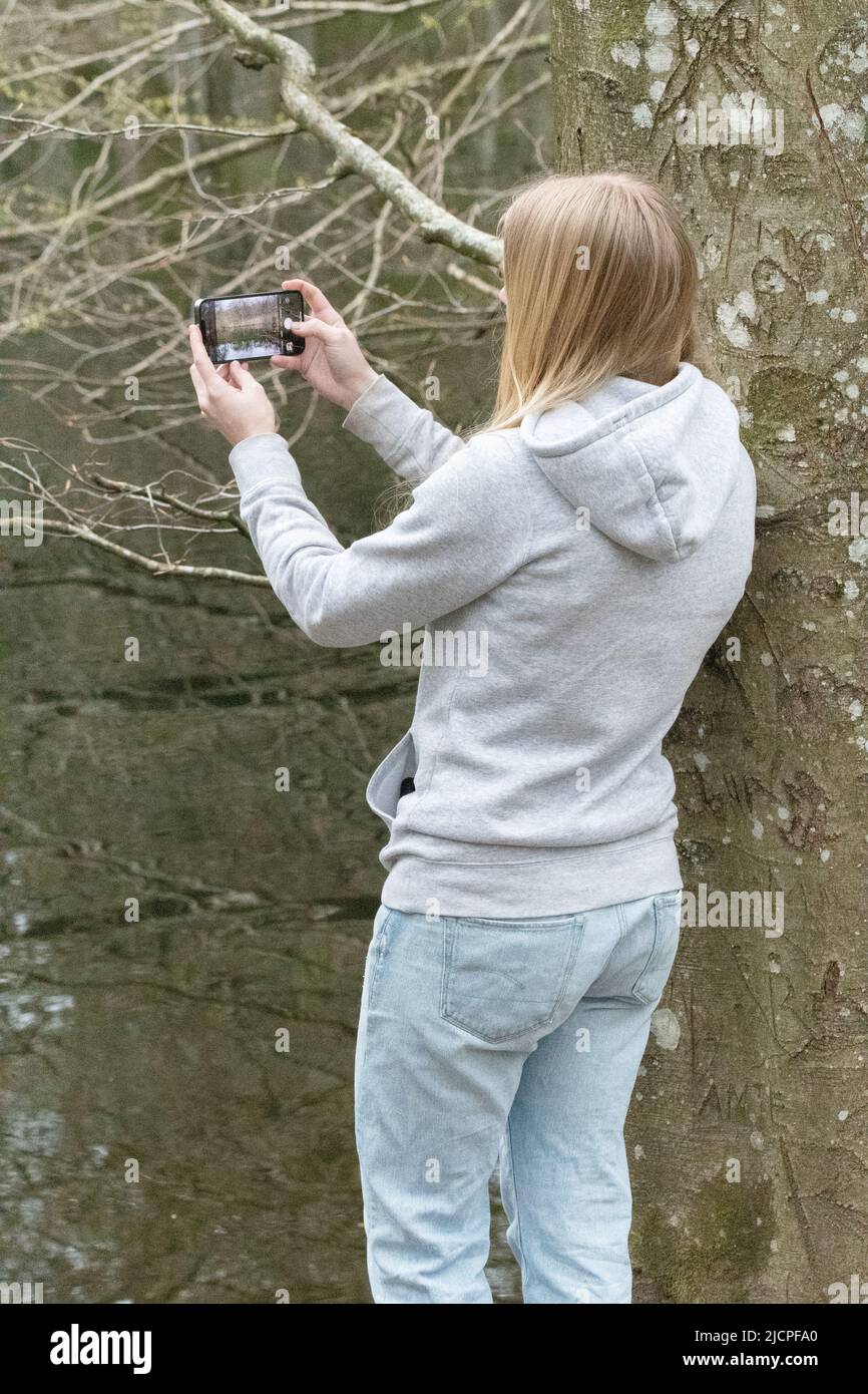 Eine junge blonde Frau von hinten, die mit ihrem Mobiltelefon an einem See fotografiert Stockfoto