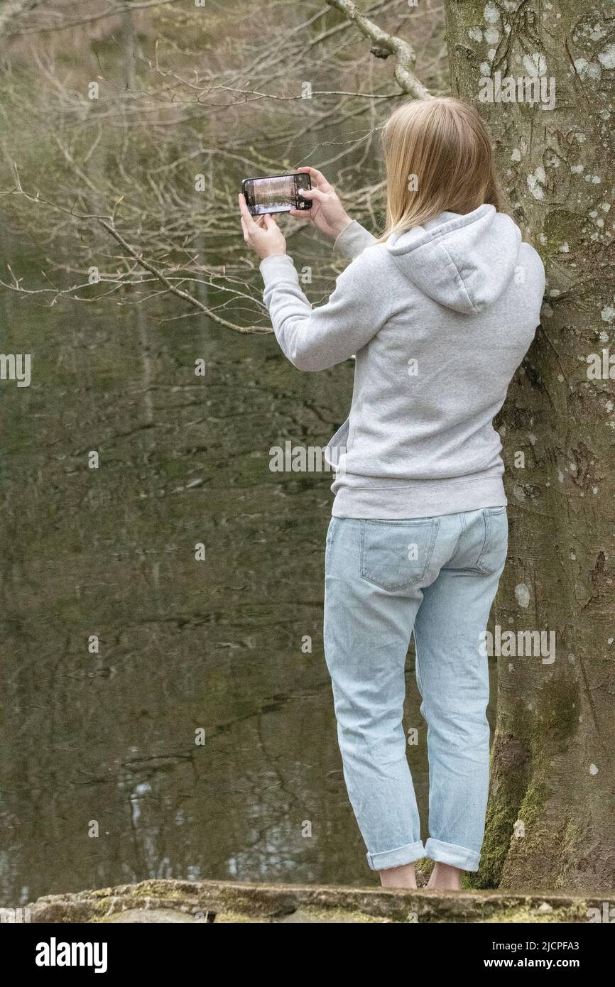 Eine junge blonde Frau von hinten, die mit ihrem Mobiltelefon an einem See fotografiert Stockfoto