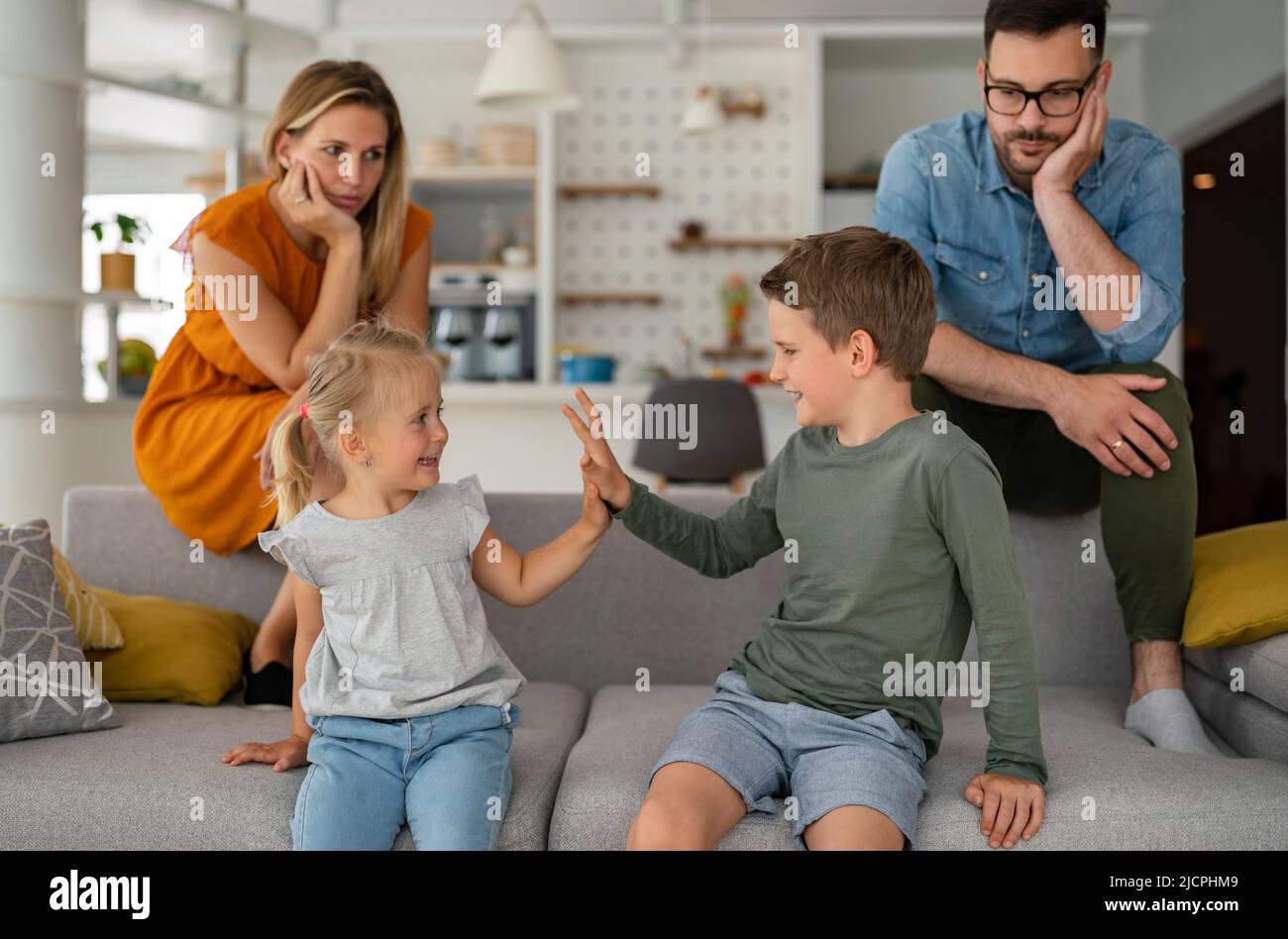 Müde Eltern, die auf der Couch sitzen, fühlen sich erschöpft genervt, während glückliche Kinder zusammen spielen. Stockfoto