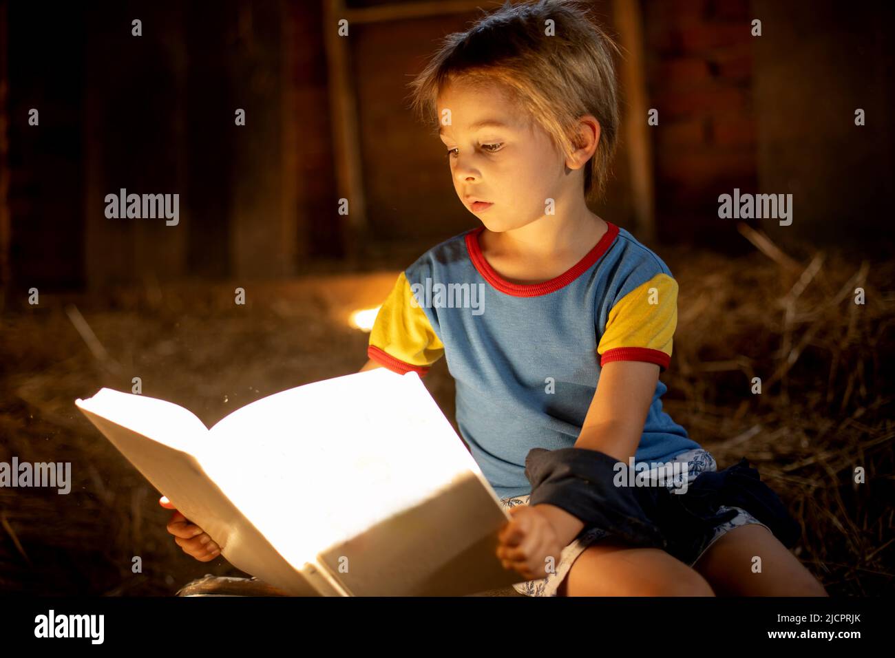 Erstaunliches, kleines blondes Kind, Junge, das ein Buch in einem mystischen, gemütlichen Raum liest, der mit Licht aus einem kleinen Fenster gefüllt ist Stockfoto