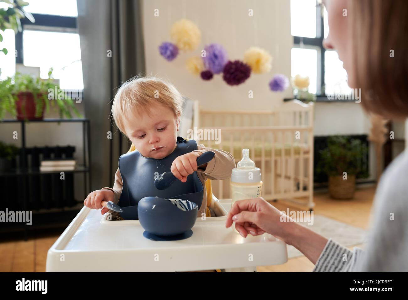 Kleines Kind im Lätzchen, das auf seinem Stuhl sitzt und lernt, mit Hilfe seiner Mutter Brei mit einem Löffel zu essen Stockfoto