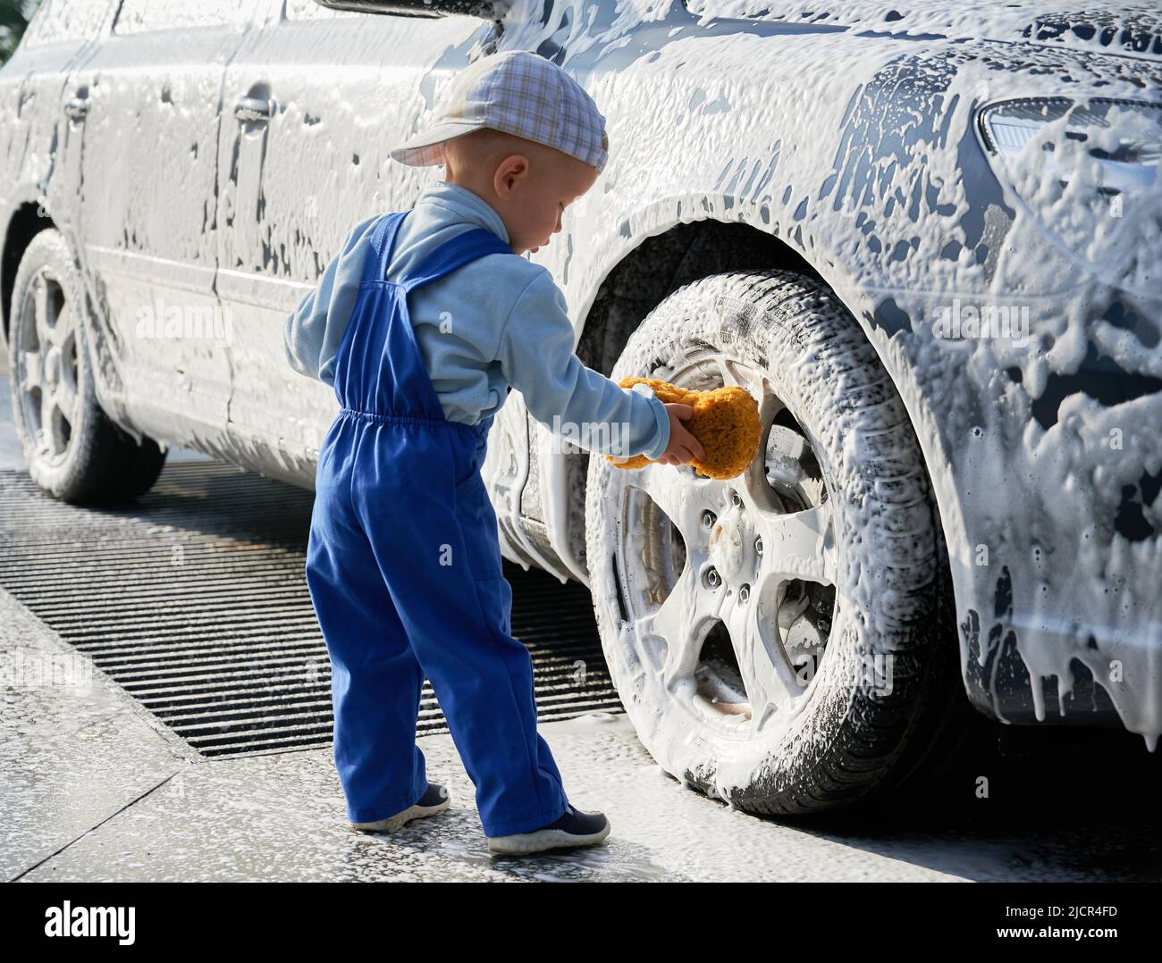Kleiner Kleinkind Junge steht in der Nähe Auto mit Schaum bedeckt, hält Schwamm in der Hand und lernen, Auto zu waschen. Männliches Kind studiert, Auto mit Schwamm zu waschen und sich bis zum Vorderrad zu beugen. Stockfoto
