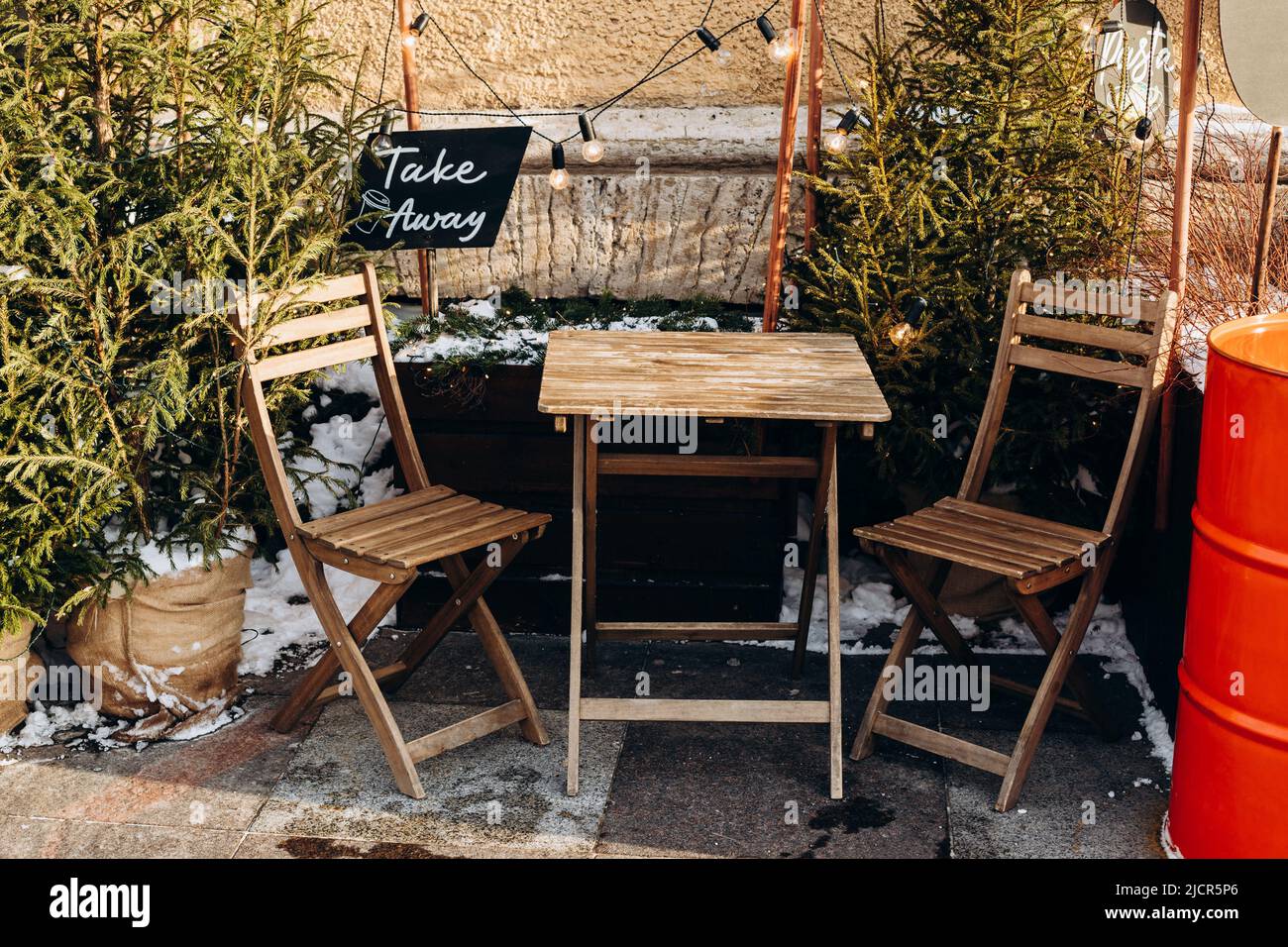 Café im Freien auf der Straße, das zu Weihnachten dekoriert und beleuchtet ist. Holztische, zwei Stühle, Tannenbäume und Schild zum Mitnehmen von Kaffee. Vorderansicht. Stockfoto Café im Freien auf der Straße, das zu Weihnachten dekoriert und beleuchtet ist. Holztische, zwei Stühle, Tannenbäume und Schild zum Mitnehmen von Kaffee. Vorderansicht. Stockfoto