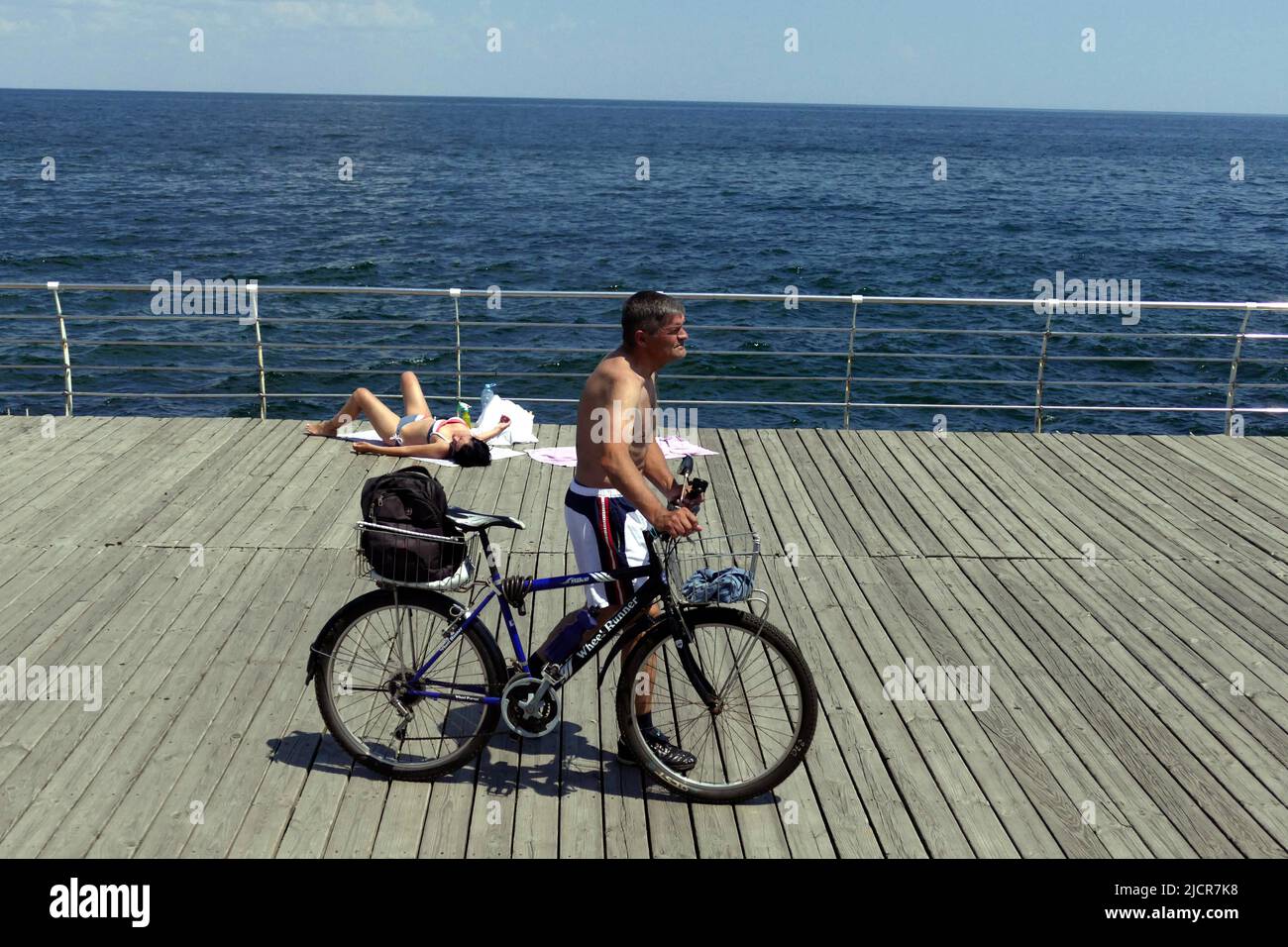 ODESA, UKRAINE - 13. JUNI 2022 - Ein Mann schiebt ein Fahrrad an einer Frau vorbei, die auf dem Holzboden des Strandes von Lanzeron bräunt, während die Anwohner prohi sind Stockfoto