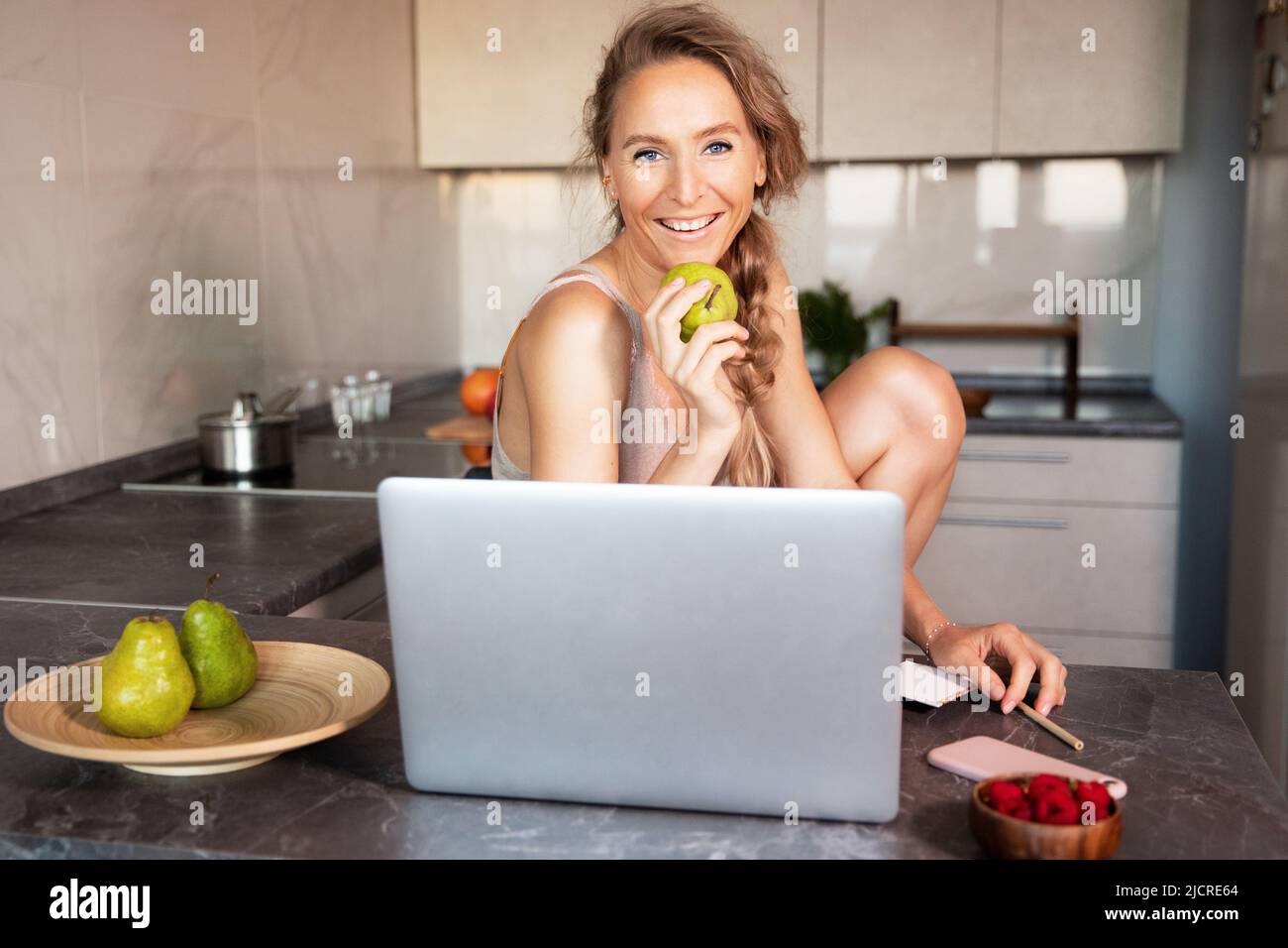 Eine junge Frau fröhlich schöne schlanke Blondine sitzt tagsüber an einem Tisch in der Küche vor einem Laptop und isst frisches Obst und Stockfoto