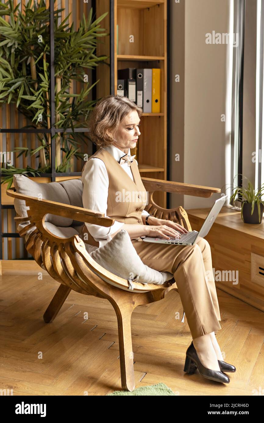 Eine ältere Geschäftsfrau in einem beigefarbenen Hosenanzug arbeitet in einem Büro, das auf einem Stuhl sitzt. Arbeiten am Computer, Tippen auf einem Laptop Stockfoto