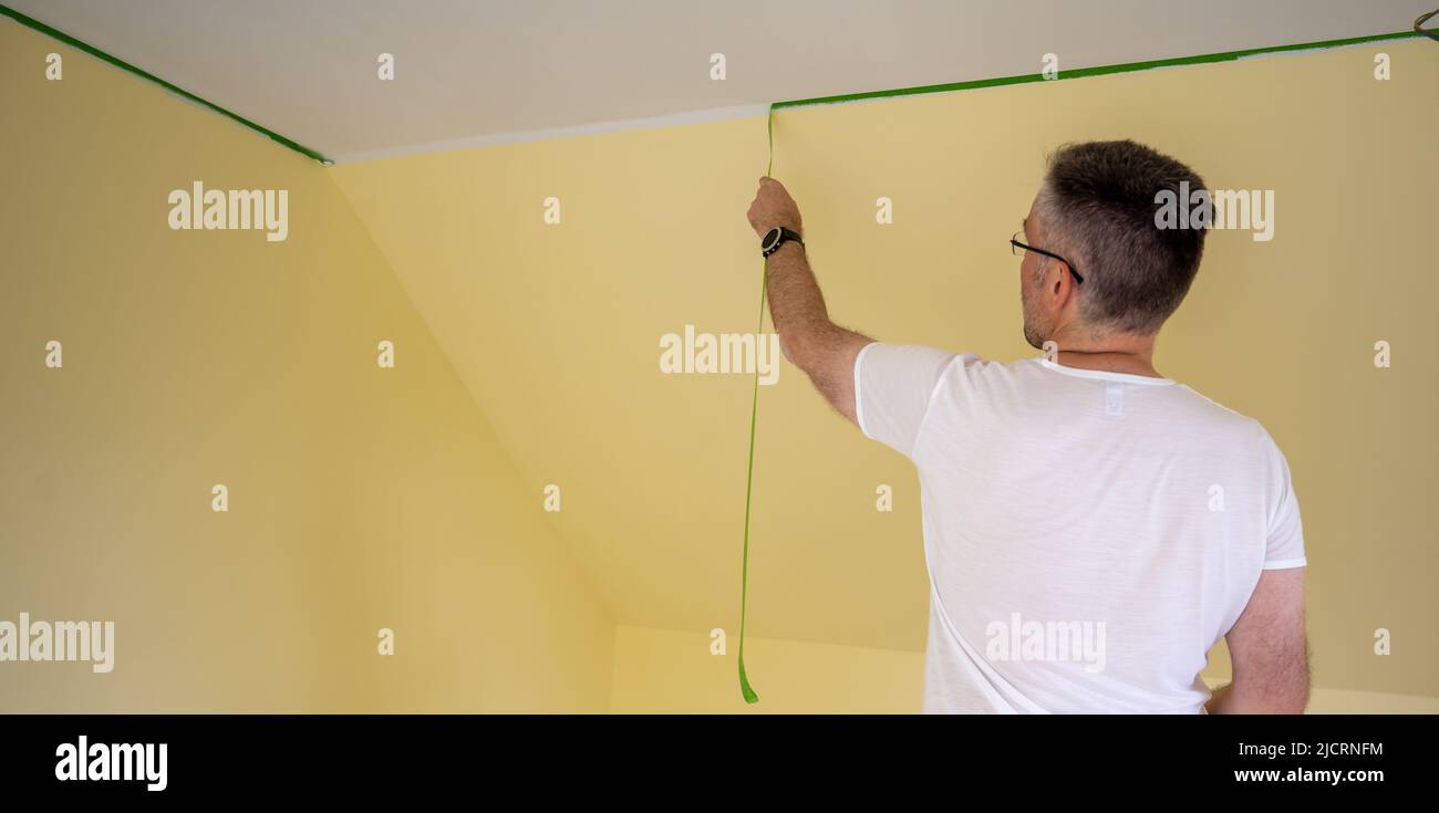 Painter entfernt Klebeband und erstellt einen scharfen Rand zwischen einem gelb-weiß lackierten Teil einer Wand. Stockfoto