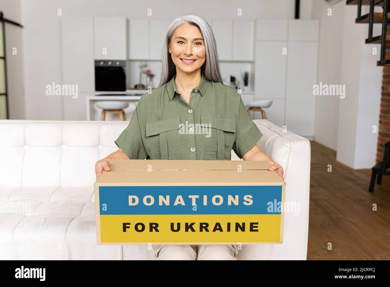 Kind smiling senior Asian female volunteer sitting on a sofa, packing goods in a cardboard box for Ukrainian people, donating to charity, worldwide people support refugees from Ukraine Stockfoto