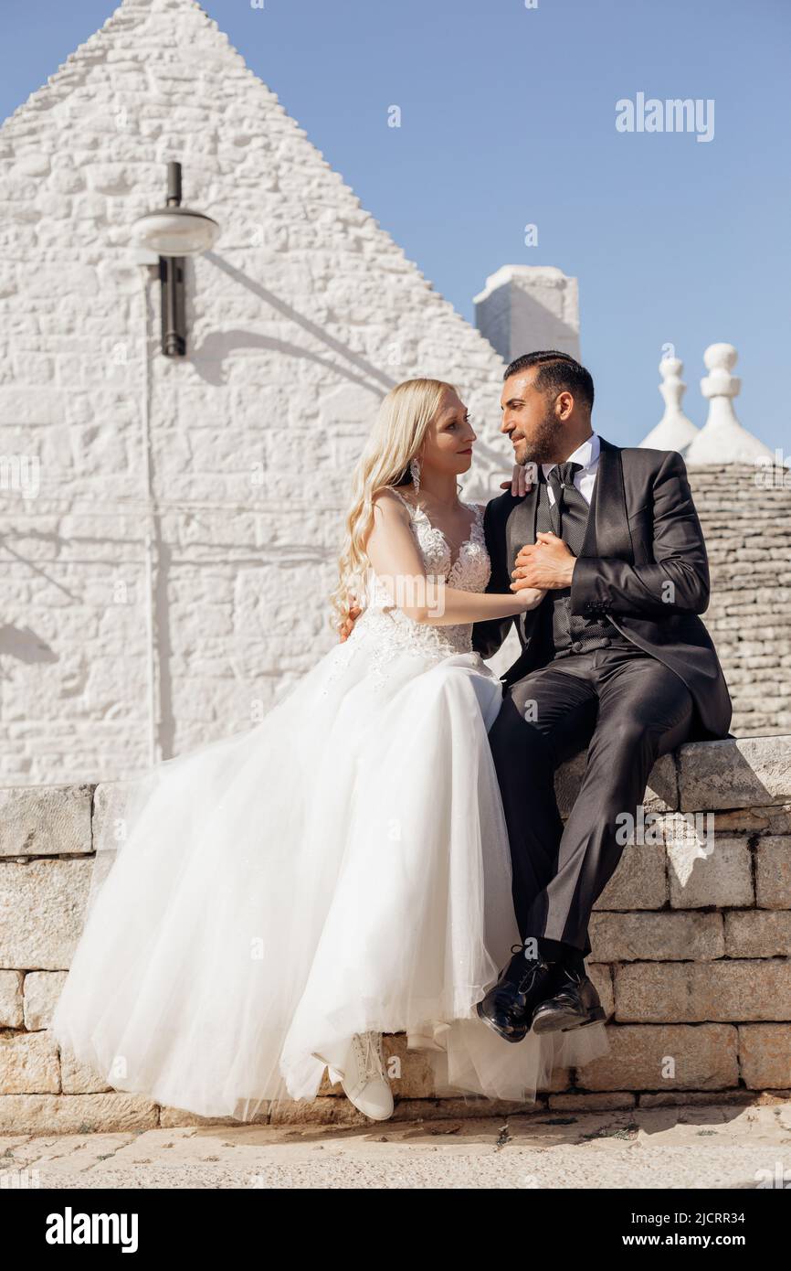 Vertikale romantische verheiratete Paar in der Liebe, blonde Frau und Mann in Anzug nach der Hochzeit, einander suchen, sitzen auf Italien Wand Stockfoto