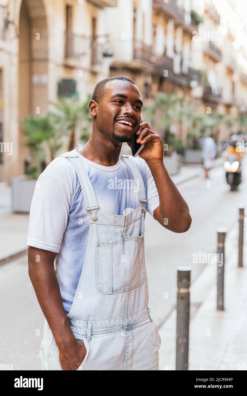 Entzückter junger afroamerikanischer stilvoller Mann in T-Shirt und insgesamt lächelnd. Er schaut weg, während er auf dem Smartphone auf der Stadtstraße mit der Hand in der Tasche steht Stockfoto