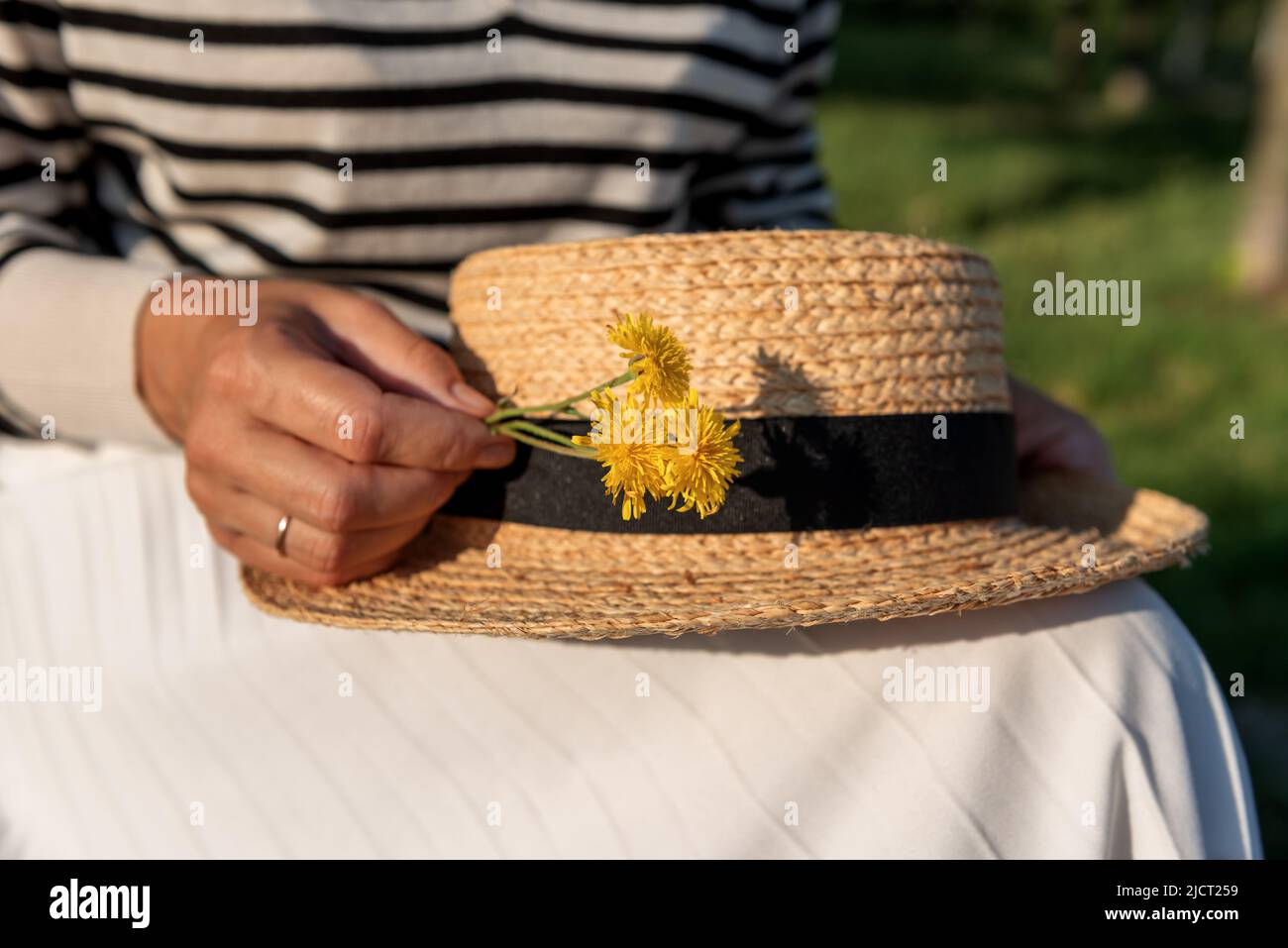 Frau in einem gestreiften Pullover und weißem Rock, die in einem Park sitzt, einen Strohhut und einen kleinen Strauß gelber Blumen auf den Knien hält, in der Mitte geschossen. Stockfoto