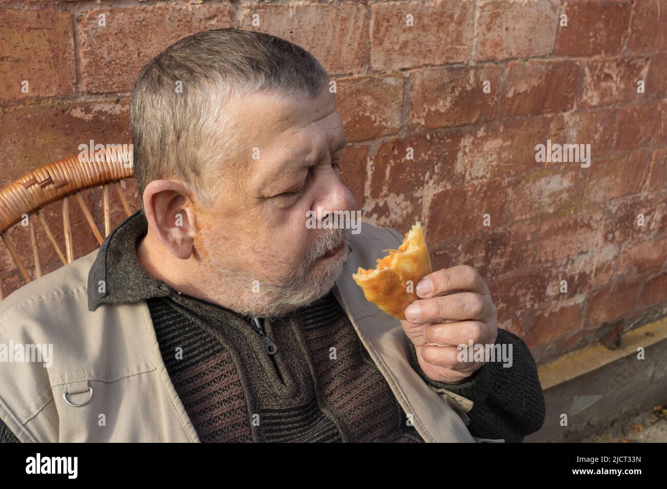 Porträt eines Ukainian Senior sitzt agaistische Backsteinmauer und essen Patty mit gebratenem Kohl gefüllt Stockfoto